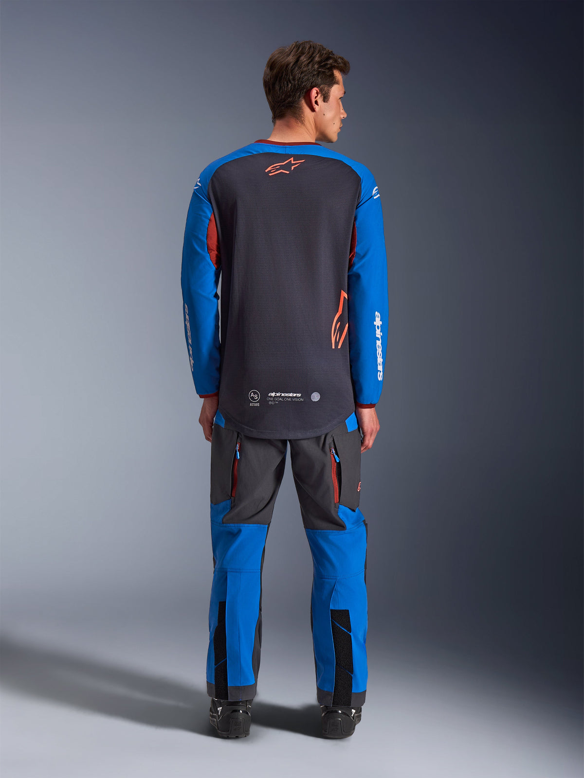Alpinestars Maxdura Dual Jersey en gris foncé et bleu roi, vue de dos montrant le torse entièrement en maille et le logo étoile orange, manches bleu roi avec marquage blanc, porté avec un pantalon d'aventure assorti, équipement haute performance pour la conduite dual-sport