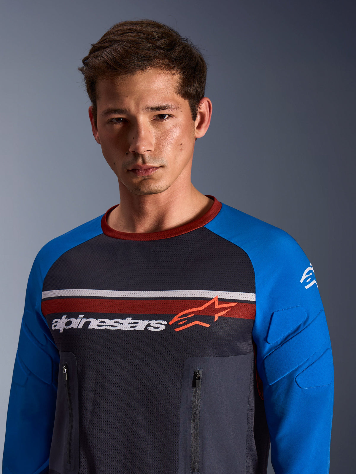 Alpinestars Maxdura Dual Jersey, gris foncé et bleu roi, porté par un mannequin homme, doté d'un torse en maille, de manches bleu roi avec rembourrage intégré et de poches poitrine zippées verticales avec marquage Alpinestars sur la poitrine.