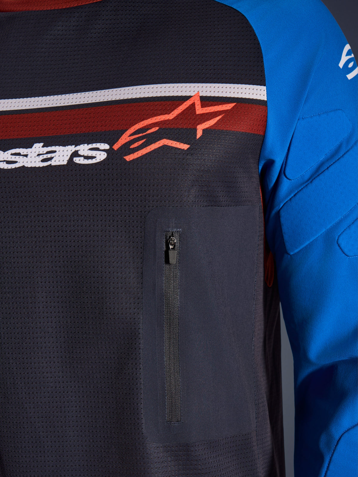 Maillot Alpinestars Maxdura Dual, gros plan sur la poitrine et la manche, gris foncé et bleu royal, avec poche poitrine zippée, torse en maille respirante et rembourrage intégré sur les manches, logo Alpinestars sur bandes rouges et blanches