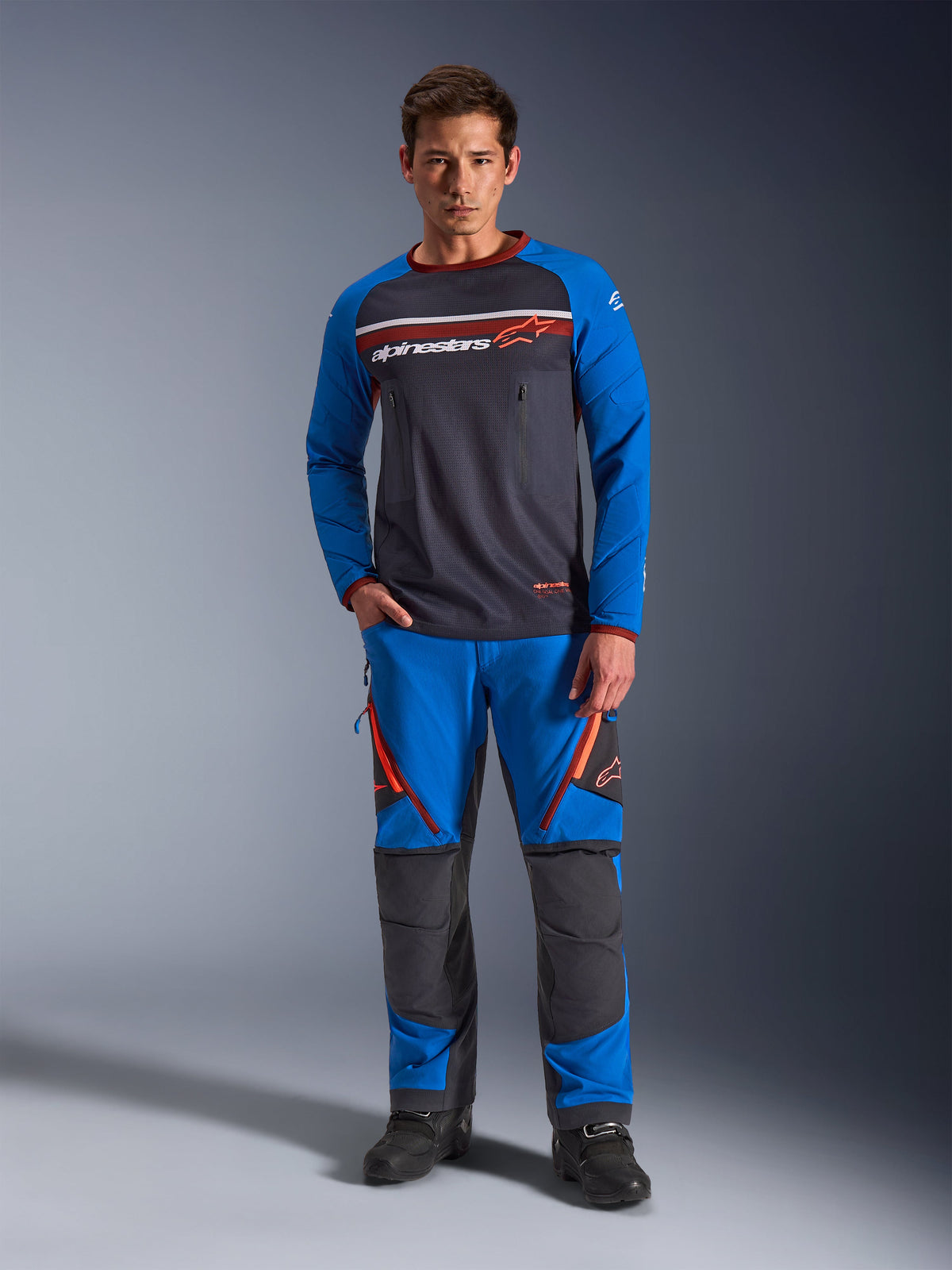 Alpinestars Maxdura Dual Jersey, maillot de moto, gris foncé et bleu royal, torse en maille intégrale avec manches rembourrées et poches poitrine zippées, logo Alpinestars blanc et orange, porté avec un pantalon d'aventure assorti et des bottes noires