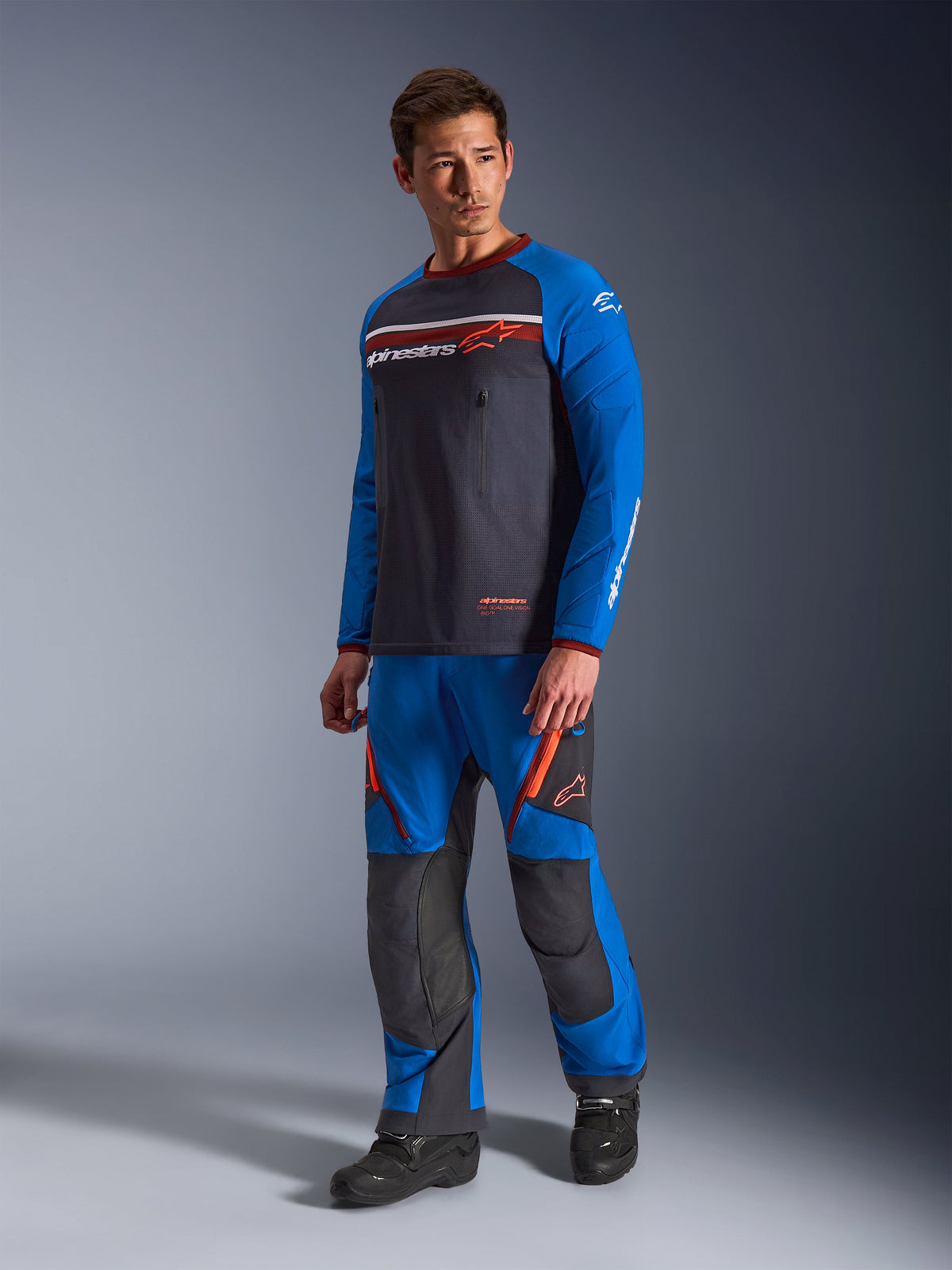 Maillot Alpinestars Maxdura Dual, gris foncé et bleu royal, maillot de moto d'aventure avec torse en maille et manches bleu royal, porté par un mannequin avec un pantalon tout-terrain assorti, doté de poches poitrine zippées et du logo Alpinestars.