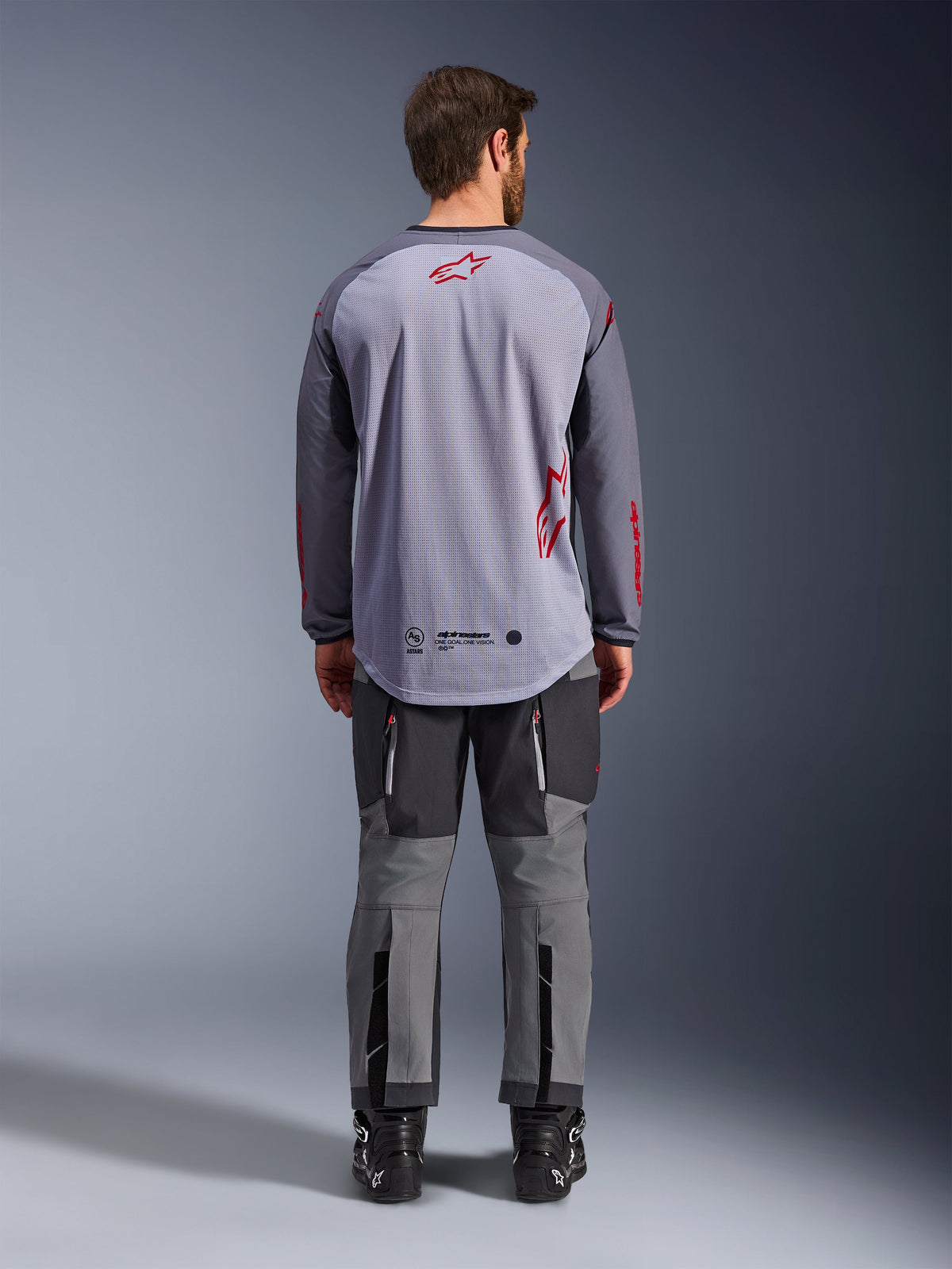 Modèle masculin vu de dos portant le maillot Alpinestars Maxdura Dual, maillot d'aventure gris moyen et gris foncé avec des accents de logo rouges, structure du dos en maille, porté avec un pantalon tout-terrain gris assorti et des bottes noires