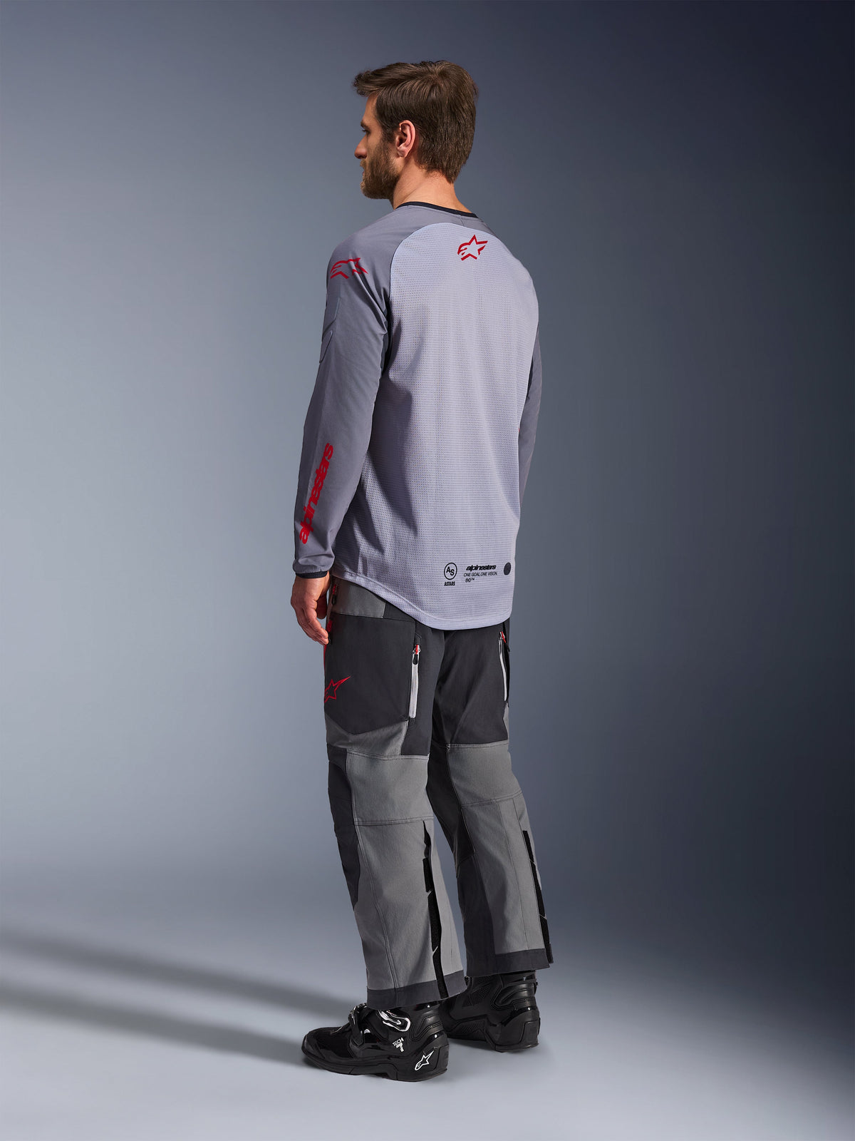 Homme portant le maillot Alpinestars Maxdura Dual, gris clair avec logos Alpinestars rouges, vue de dos montrant le torse en maille et les manches longues, associé à un pantalon technique anthracite et des bottes de moto noires