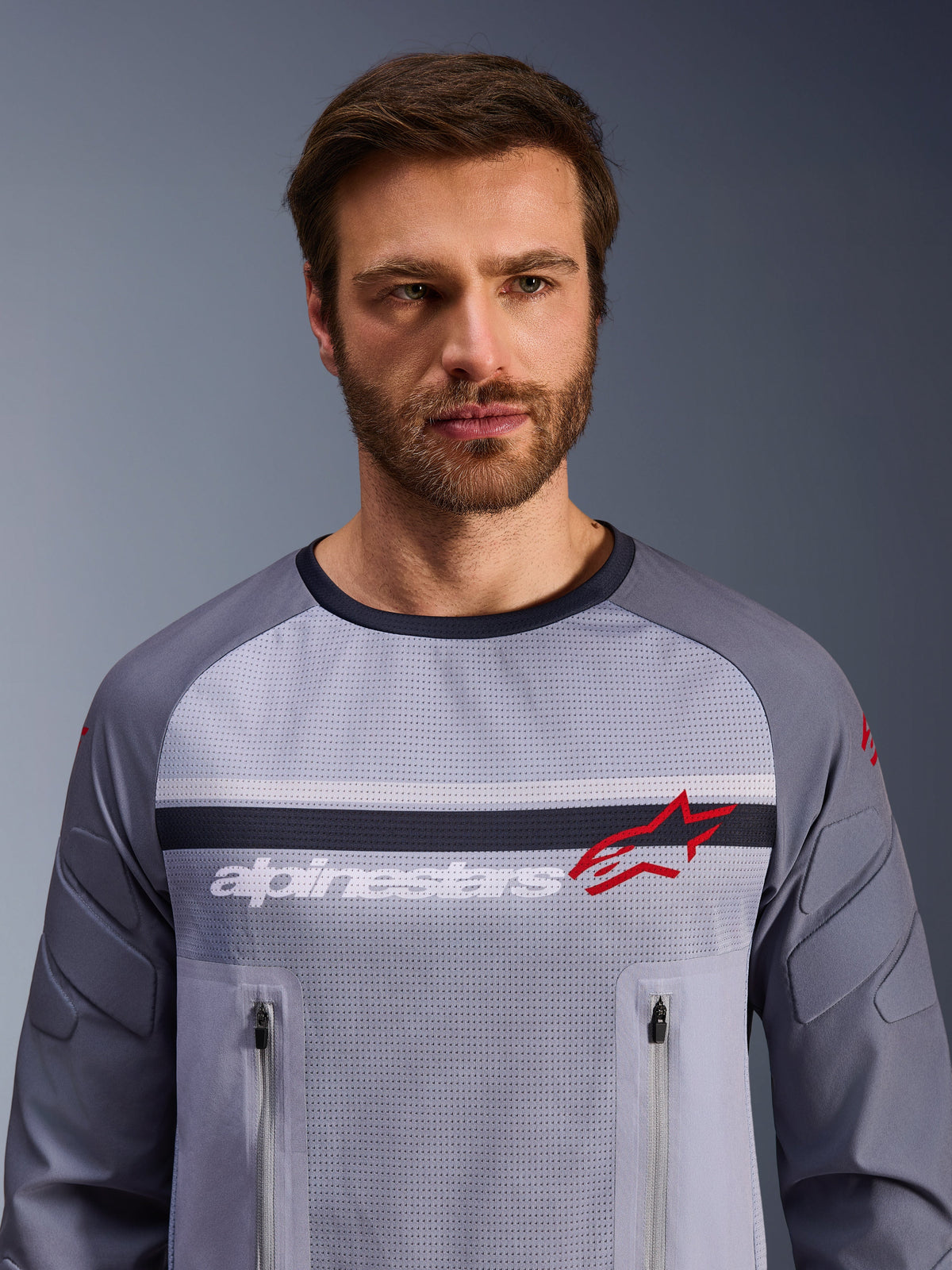 Maillot Alpinestars Maxdura Dual, gris moyen et gris foncé, porté par un mannequin homme, doté d'un torse en maille pour la ventilation, deux poches poitrine zippées, marquage 'alpinestars' blanc et logo étoile rouge, équipement de performance pour l'aventure tout-terrain