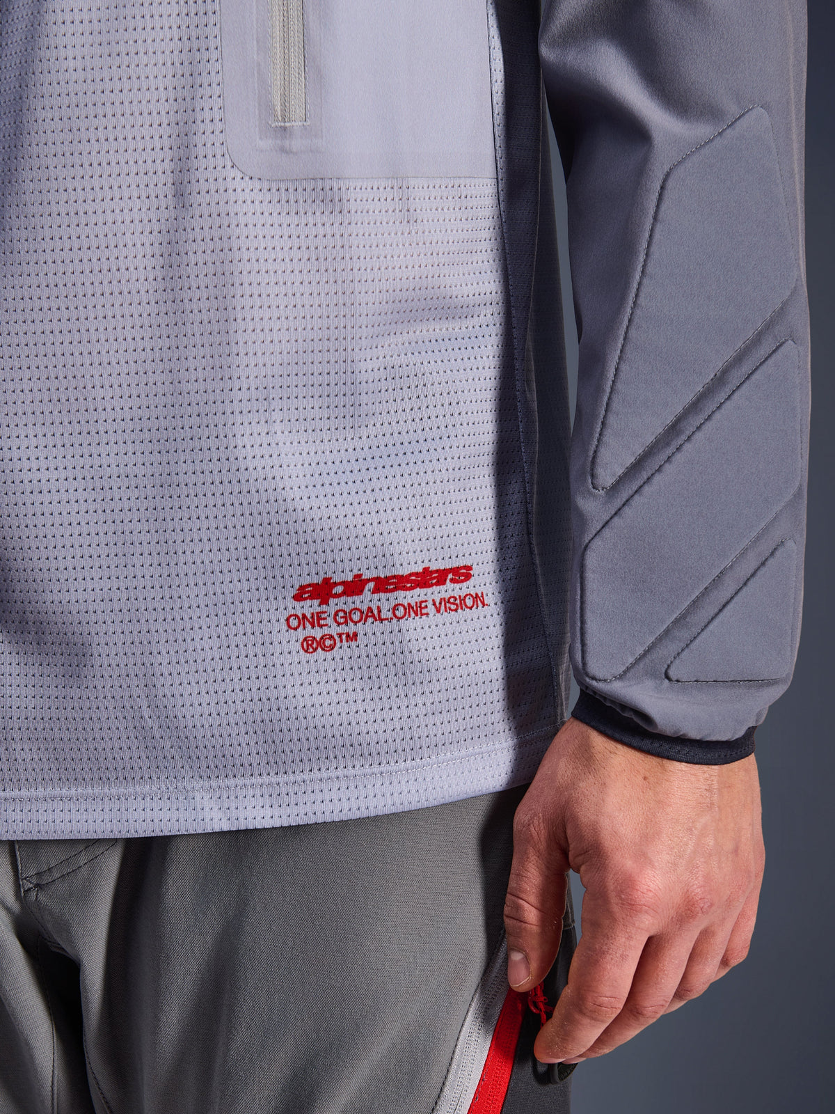 Maillot Alpinestars Maxdura Dual, gris moyen et gris foncé, gros plan montrant le torse en maille respirante avec logo rouge, détail de la poche zippée et rembourrage intégré sur la manche, équipement technique de moto d'aventure