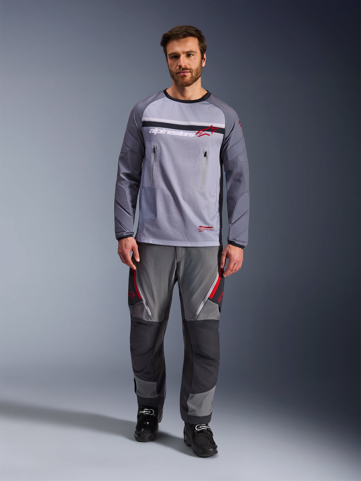 Maillot Alpinestars Maxdura Dual, gris moyen et gris foncé, maillot de moto d'aventure avec torse en maille et poches poitrine zippées, manches résistantes à l'abrasion avec rembourrage intégré, porté avec un pantalon de performance gris assorti avec des accents rouges, équipement technique de conduite tout-terrain