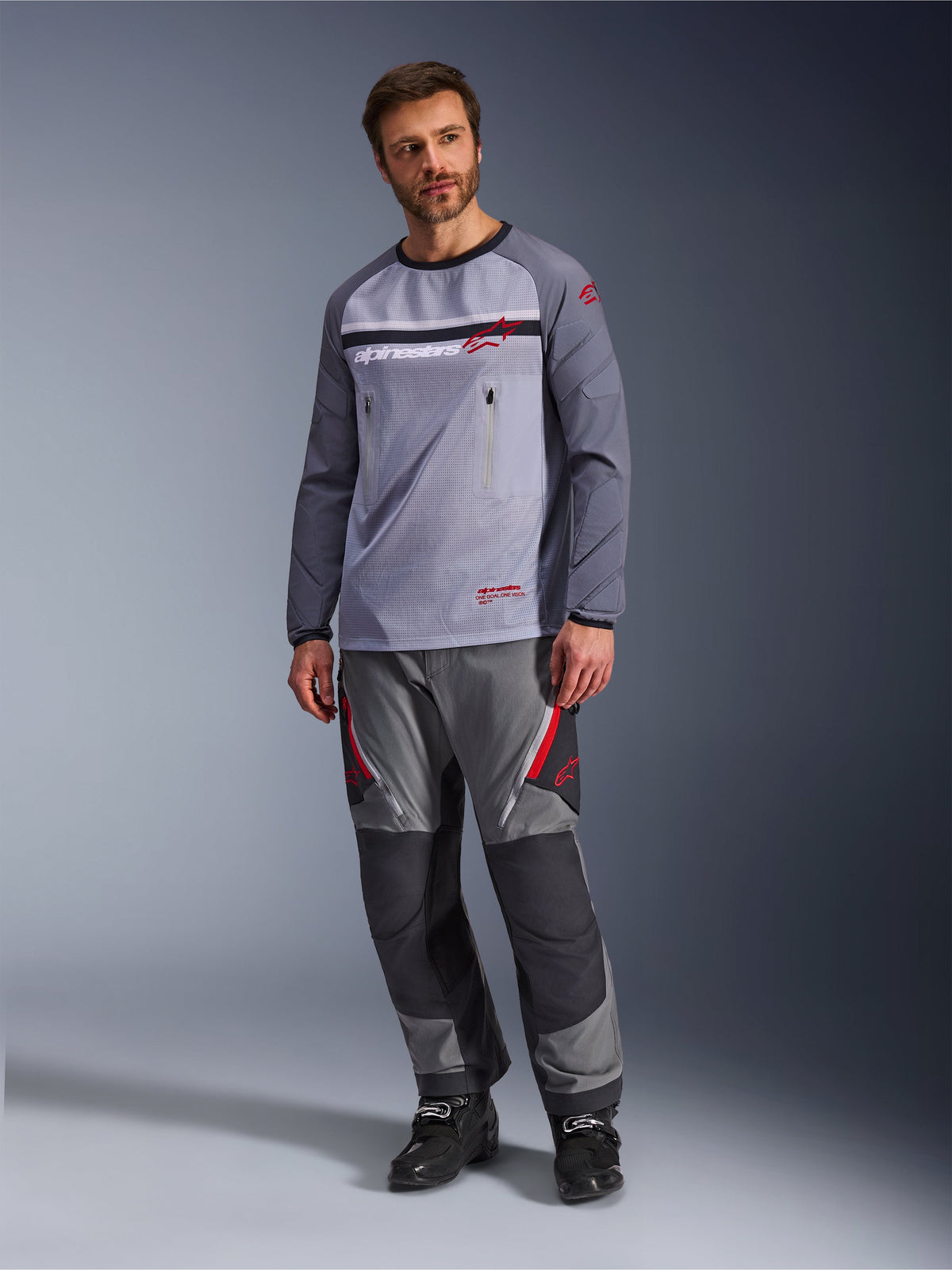 Homme portant le maillot Alpinestars Maxdura Dual en Mid Gray et Dark Gray, avec pantalon d'aventure gris et noir coordonné et bottes noires, torse en maille respirante, poches poitrine zippées et accents de logo rouges.
