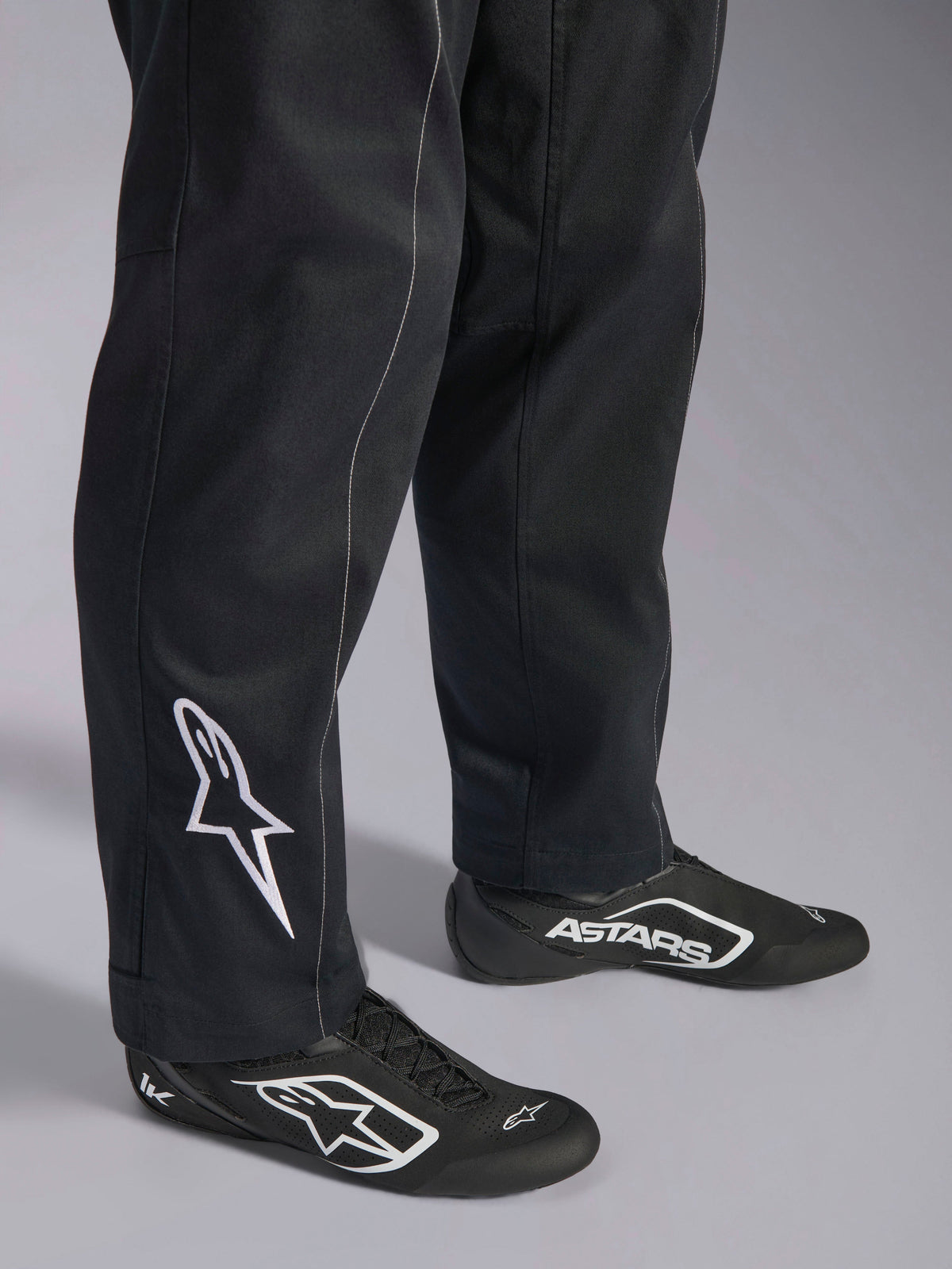 Combinaison Alpinestars Kart Indoor V2, combinaison de karting noire, gros plan sur le bas des jambes et les pieds avec un logo Alpinestars blanc sur le mollet, portée avec des chaussures de course assorties noires et blanches, conçue pour le karting de loisir
