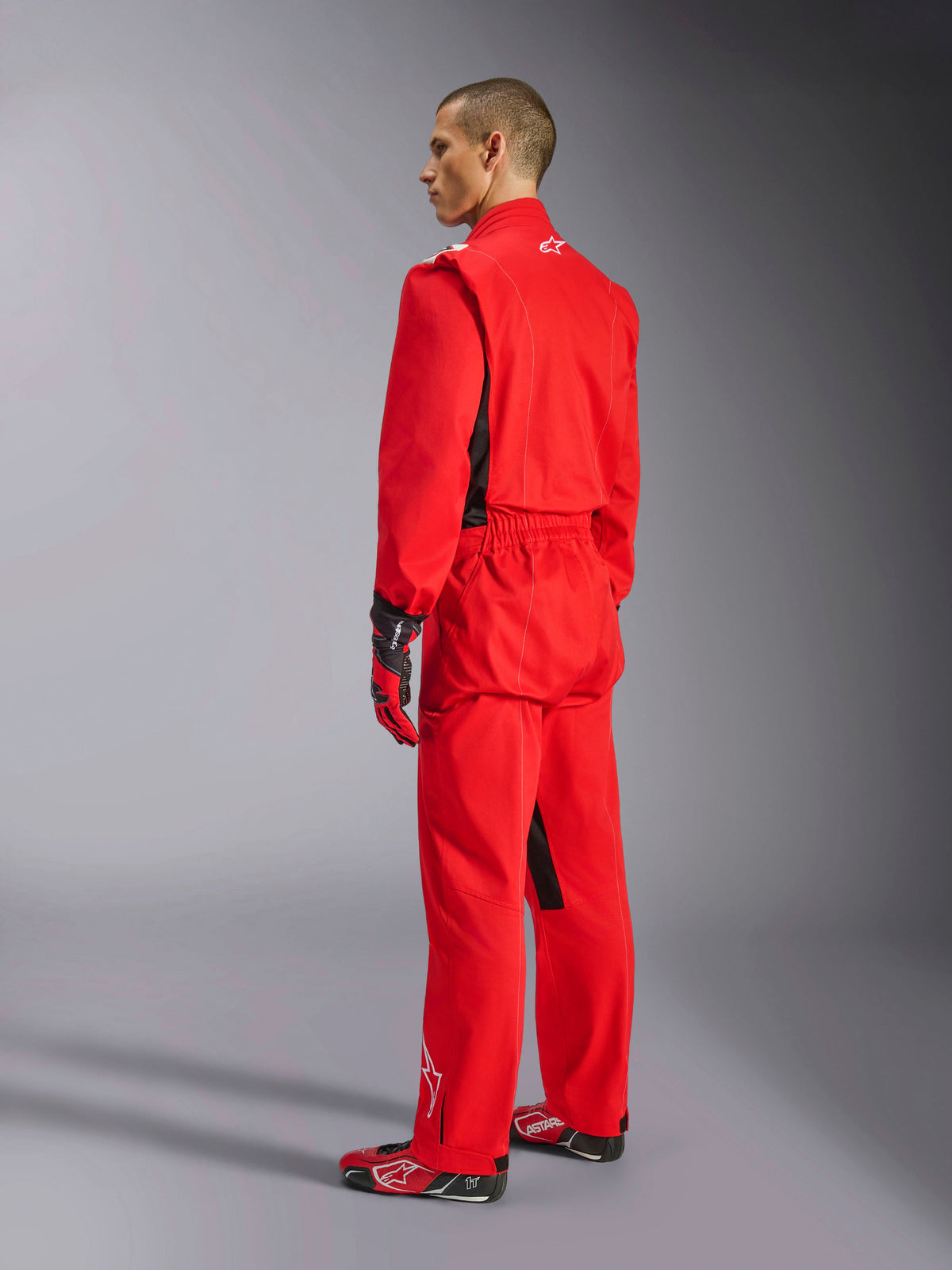 Alpinestars Kart Indoor V2 Anzug, roter Kart-Anzug, Dreiviertel-Rückansicht an einem Modell, getragen mit passenden roten Handschuhen und Rennschuhen, einlagige Poly-Baumwoll-Twill-Konstruktion, professionelle Motorsportausrüstung für Indoor-Kartfahren.