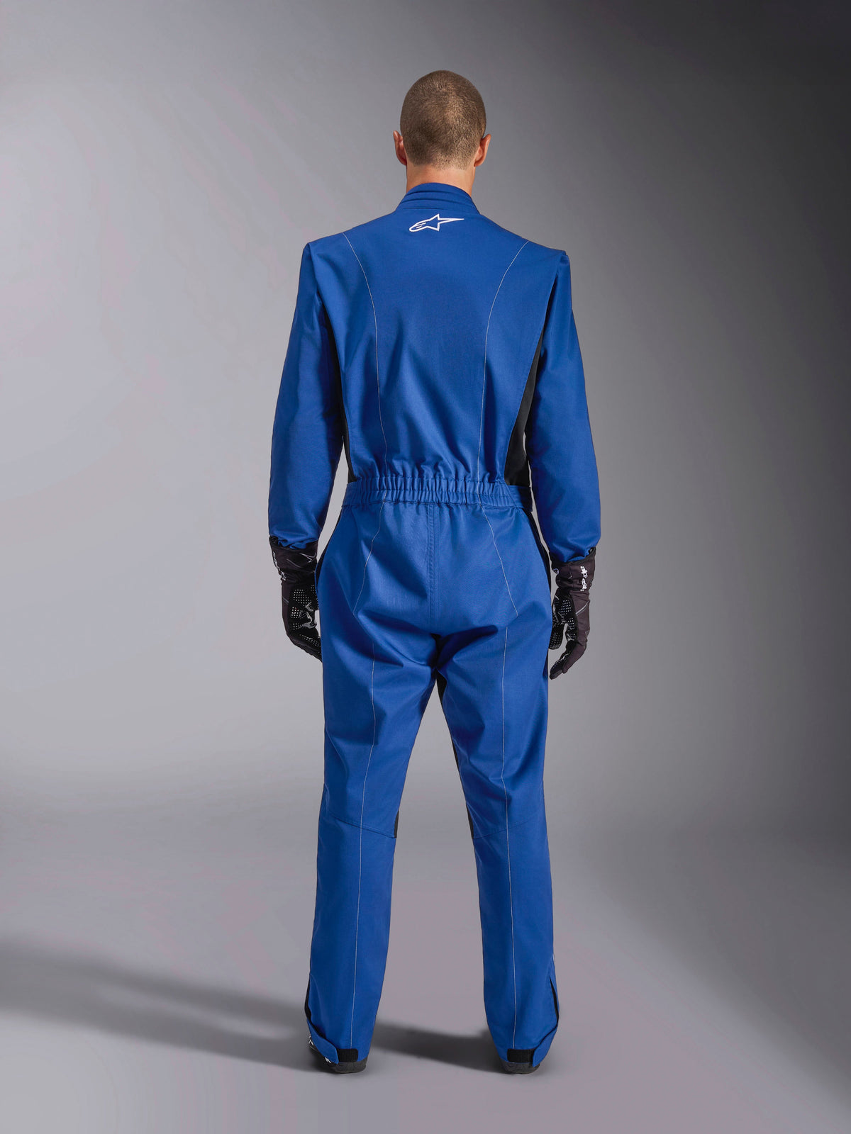 Alpinestars Kart Indoor V2 Anzug, Blau, Rückansicht, einteiliger Kart-Anzug mit weißem Logo auf dem oberen Rücken, elastischem Taillenbund und schwarzen Akzenten, getragen mit schwarzen Rennhandschuhen