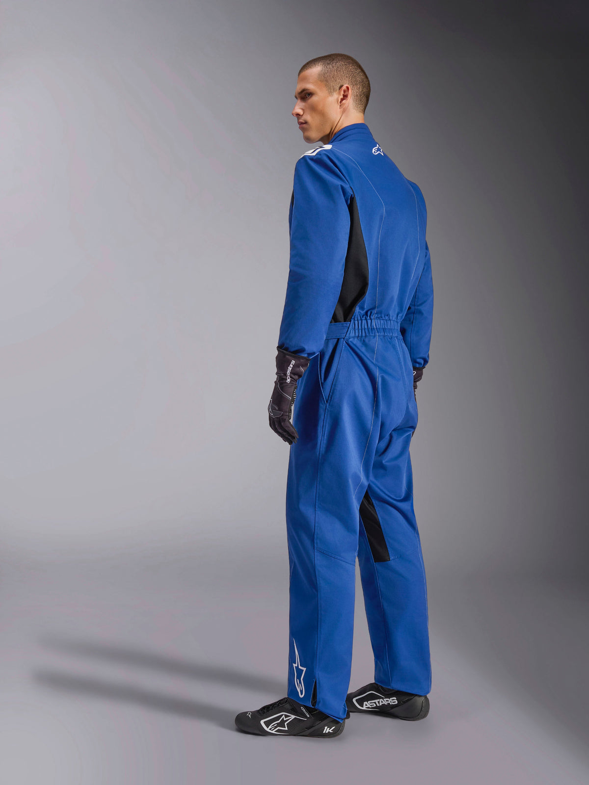 Alpinestars Kart Indoor V2 Anzug, blauer Kart-Anzug, Rück- und Seitenansicht, einteiliger Rennanzug mit schwarzen Seiteneinsätzen und Akzenten an den Beininnenseiten, elastischer Bund und weiße Logos, kombiniert mit schwarzen Rennhandschuhen und Rennschuhen