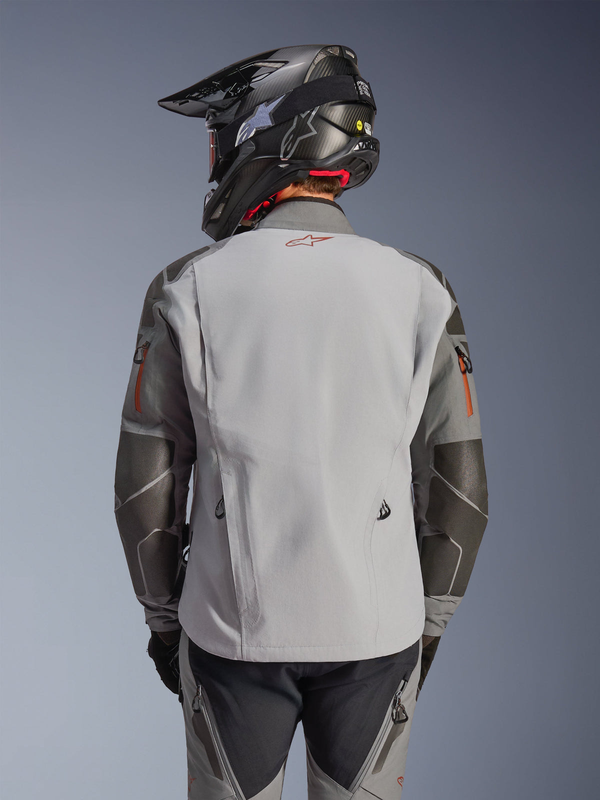 Alpinestars Nazca 3L GORE-TEX PRO Jacket, Adventure Motorcycle Jacket, Back View, Dark Gray and Mid Gray, features Superfabric reinforcements on elbows and shoulders, red logo on the back, worn with matching adventure gear and a carbon fiber helmet.