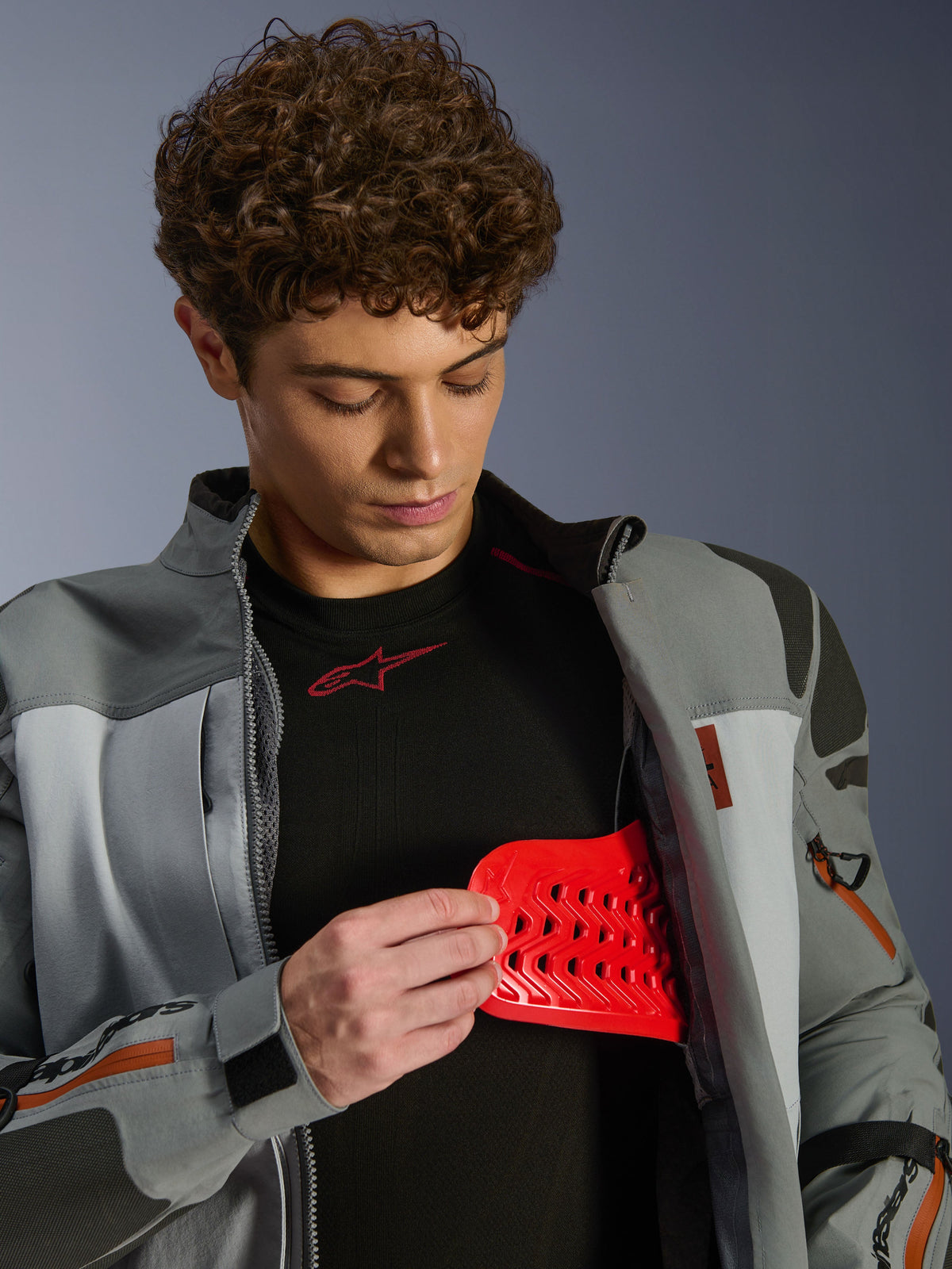 Male model wearing Alpinestars Nazca 3L GORE-TEX PRO Jacket, Dark Gray and Mid Gray, displaying red Nucleon PLASMA Pro armor insert, adventure riding apparel, premium motorcycle jacket with technical details