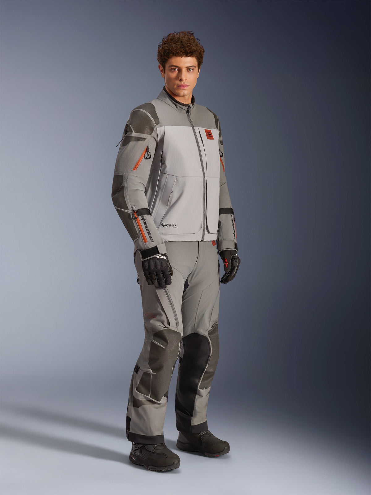 Alpinestars Nazca 3L GORE-TEX PRO Jacket in Dark Gray Mid Gray, adventure motorcycle riding outfit worn with matching pants and black gloves, featuring orange zipper accents, protective Superfabric® reinforcements, and GORE-TEX branding.