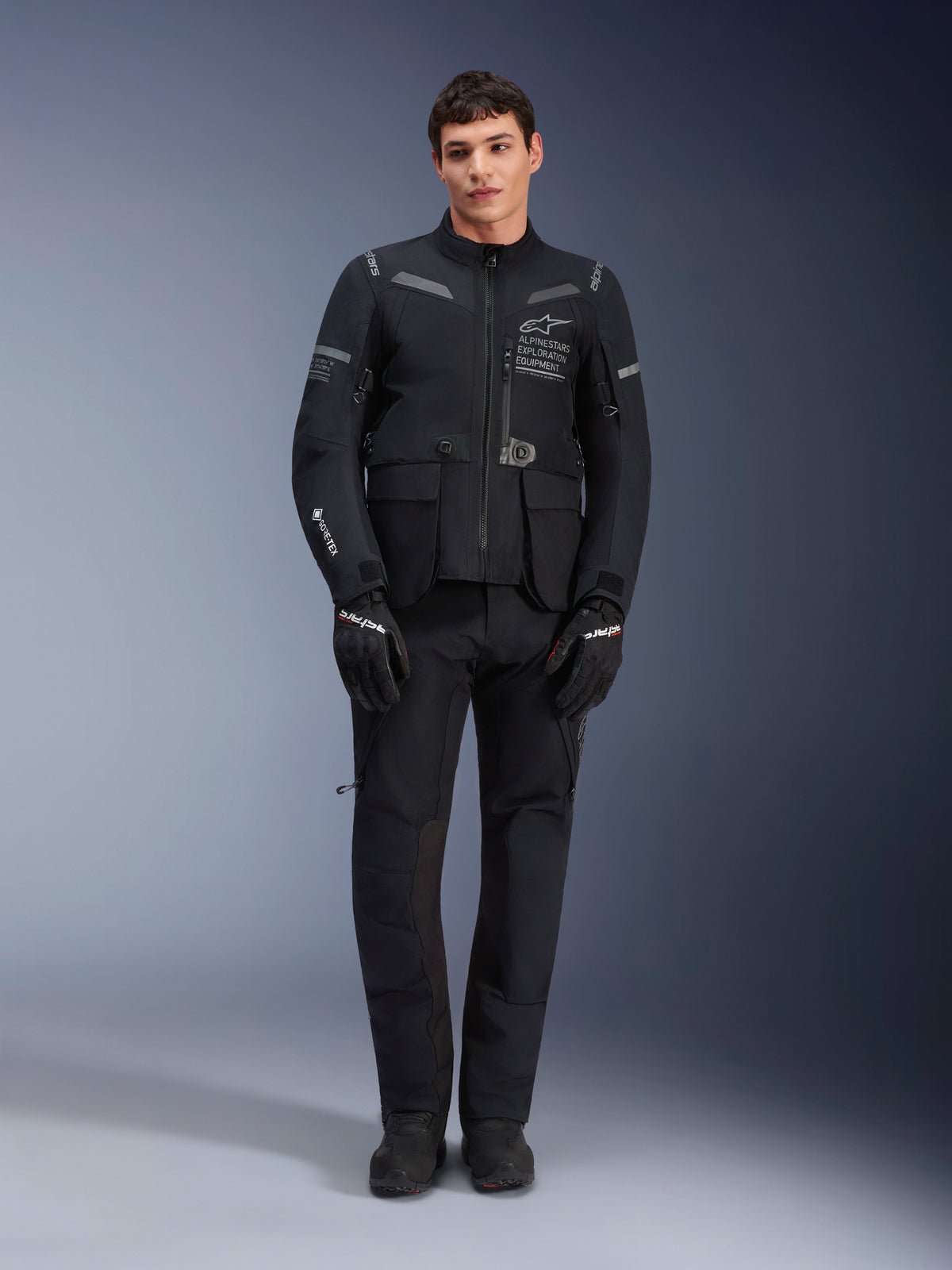 Alpinestars ST-7 2L GORE-TEX Jacke, Adventure-Touring-Motorradjacke, Schwarz und Dunkelgrau, getragen mit passender schwarzer Motorradhose und Handschuhen, mit mehreren Cargotaschen, reflektierenden Akzenten und Alpinestars Exploration Equipment-Branding auf der Brust, funktionales Design für Straßen- und Geländefahrten