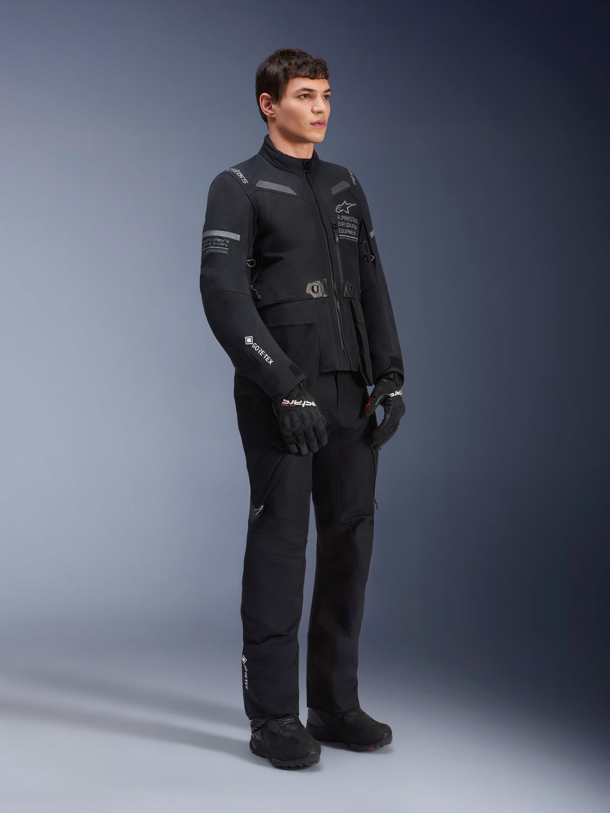 Männliches Model trägt Alpinestars ST-7 2L GORE-TEX Jacke in Black Dark Gray, Adventure-Touring-Motorradjacke, passende schwarze Hose und Handschuhe, Dreiviertel-Ganzkörperansicht, mit GORE-TEX Logo und Alpinestars Exploration Equipment Branding
