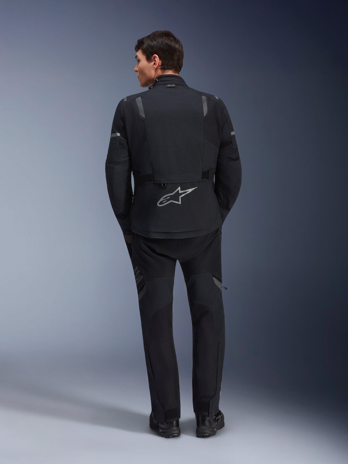 Veste Alpinestars ST-7 2L GORE-TEX et pantalon ST-7 2L GORE-TEX assorti, équipement de moto d'aventure noir et gris foncé, vue arrière montrant le logo Alpinestars réfléchissant sur la veste et la construction textile technique.