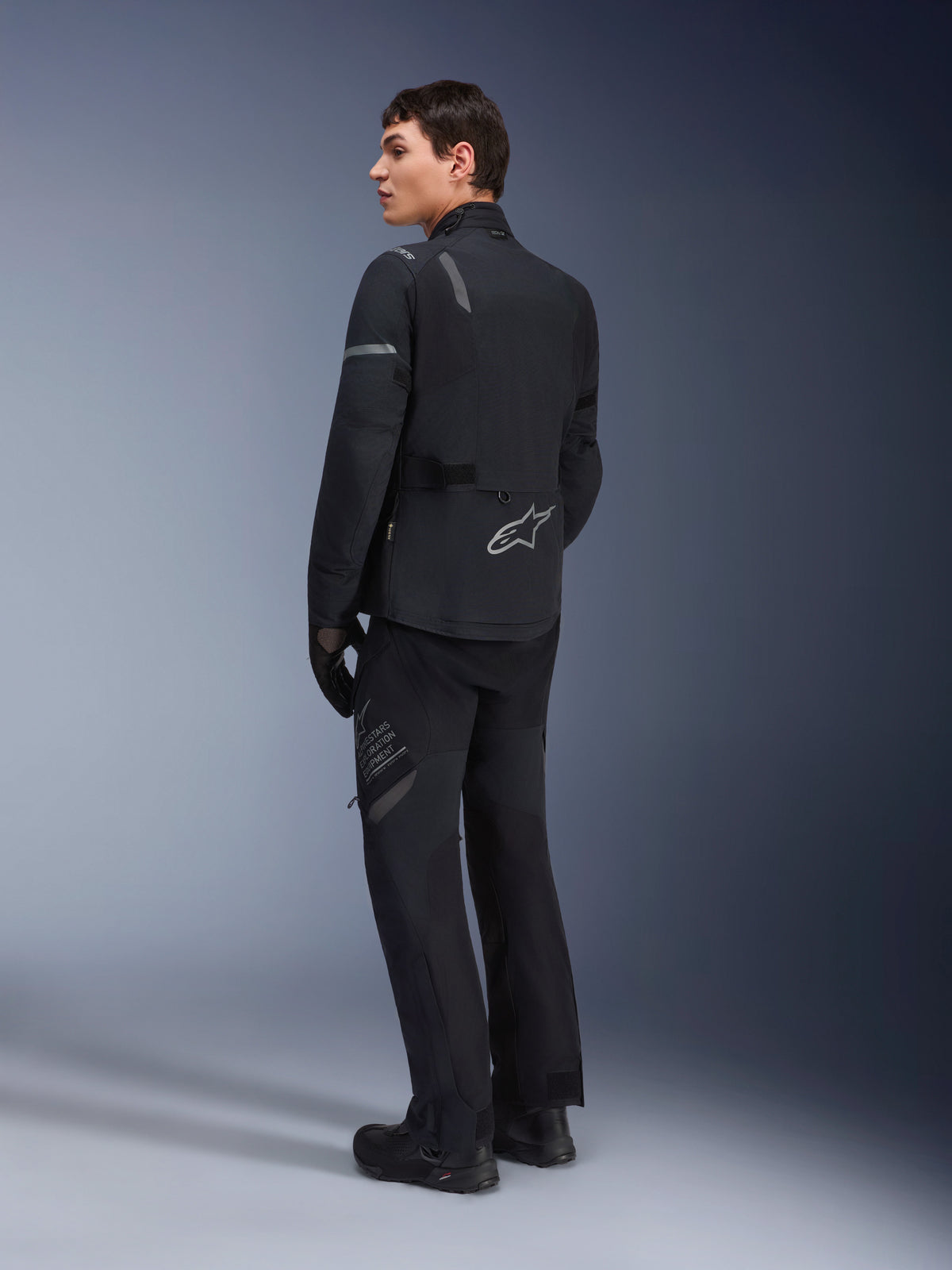 Männliches Model trägt die Alpinestars ST-7 2L GORE-TEX Jacke in Schwarz-Dunkelgrau, Dreiviertel-Rückansicht mit reflektierenden Akzenten und silbernem Logo am unteren Rücken, Adventure-Touring-Motorradbekleidung, kombiniert mit passender schwarzer Fahrhose und Stiefeln