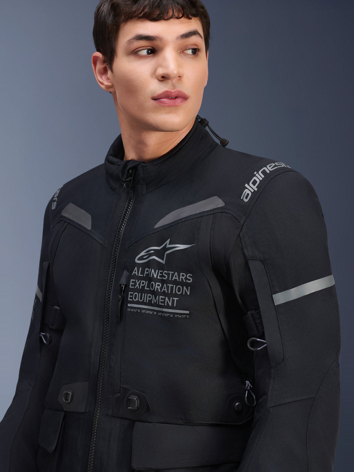 Alpinestars ST-7 2L GORE-TEX Jacke, Motorradjacke, Schwarz und Dunkelgrau, getragen von einem männlichen Model, mit Alpinestars Exploration Equipment-Branding auf der Brust, reflektierenden grauen Akzenten und abenteuertauglicher Textilkonstruktion