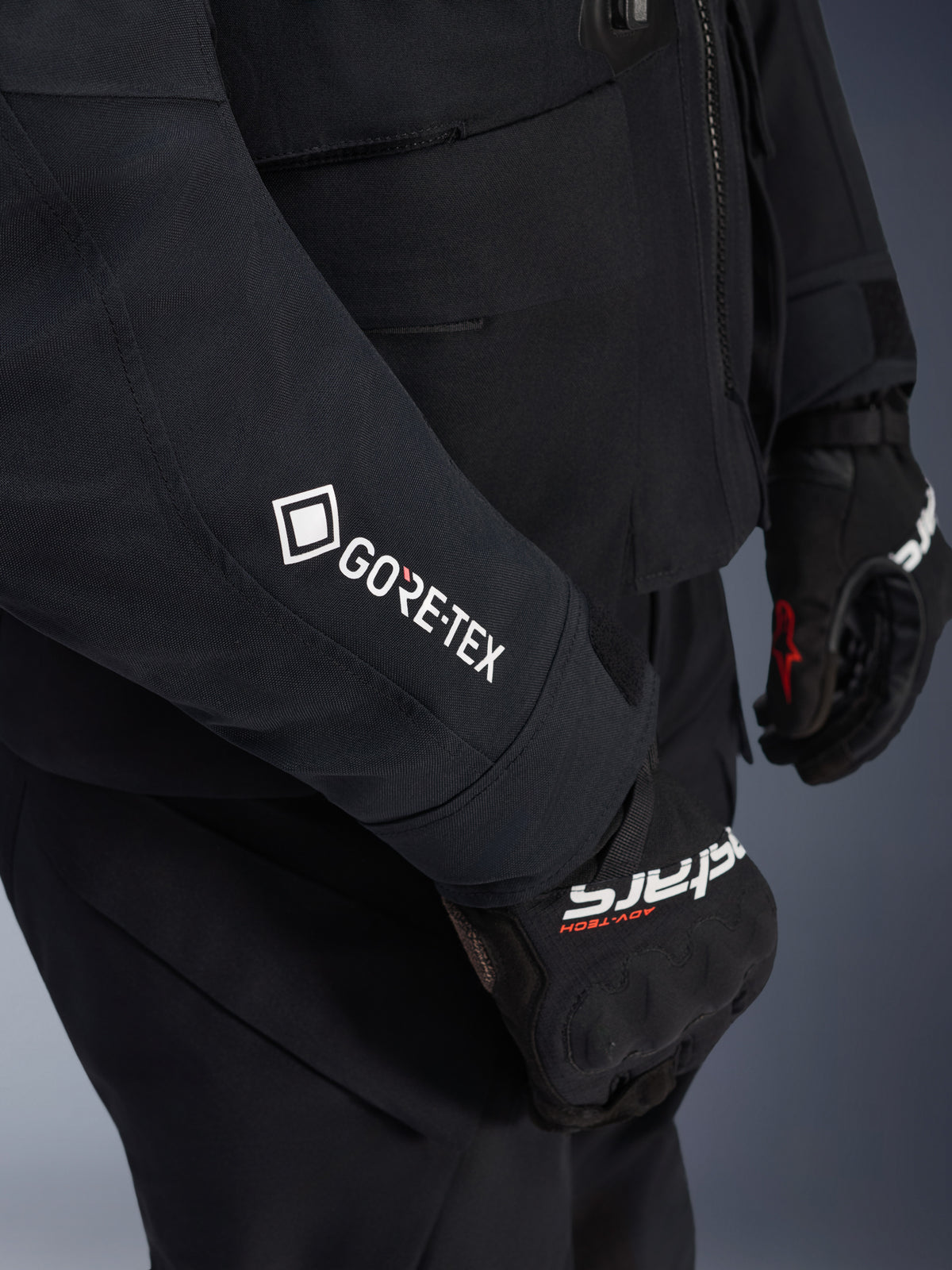 Alpinestars ST-7 2L GORE-TEX Jacke, Schwarz und Dunkelgrau, Nahaufnahme des Ärmels mit weißem GORE-TEX-Branding, getragen mit schwarzen Adventure-Motorradhandschuhen, wasserdichte Motorradbekleidung