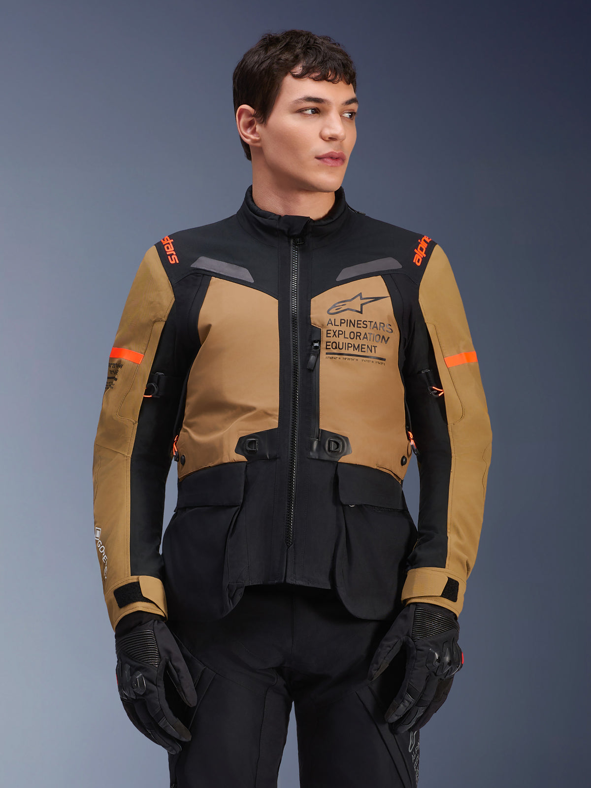 Alpinestars ST-7 2L GORE-TEX motorjas, adventure touring motorjas, Gobi Brown en zwart met oranje accenten, gedragen door een mannelijk model, voorzien van Exploration Equipment-branding en GORE-TEX-technologie, duurzame waterdichte motoruitrusting.