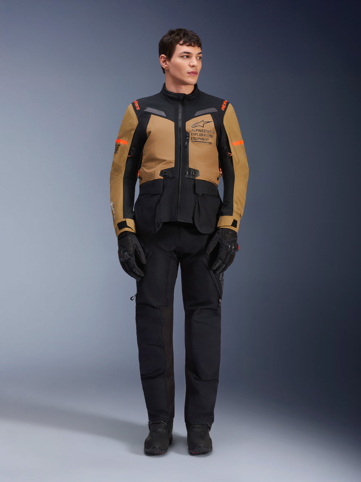 Mannelijk model draagt Alpinestars ST-7 2L GORE-TEX Jas, Gobi Brown en zwart, adventure motoruitrusting met bijpassende zwarte broek en handschoenen, voorzien van Alpinestars Exploration Equipment-branding op de borst, functioneel waterdicht ontwerp voor road- en off-road touring