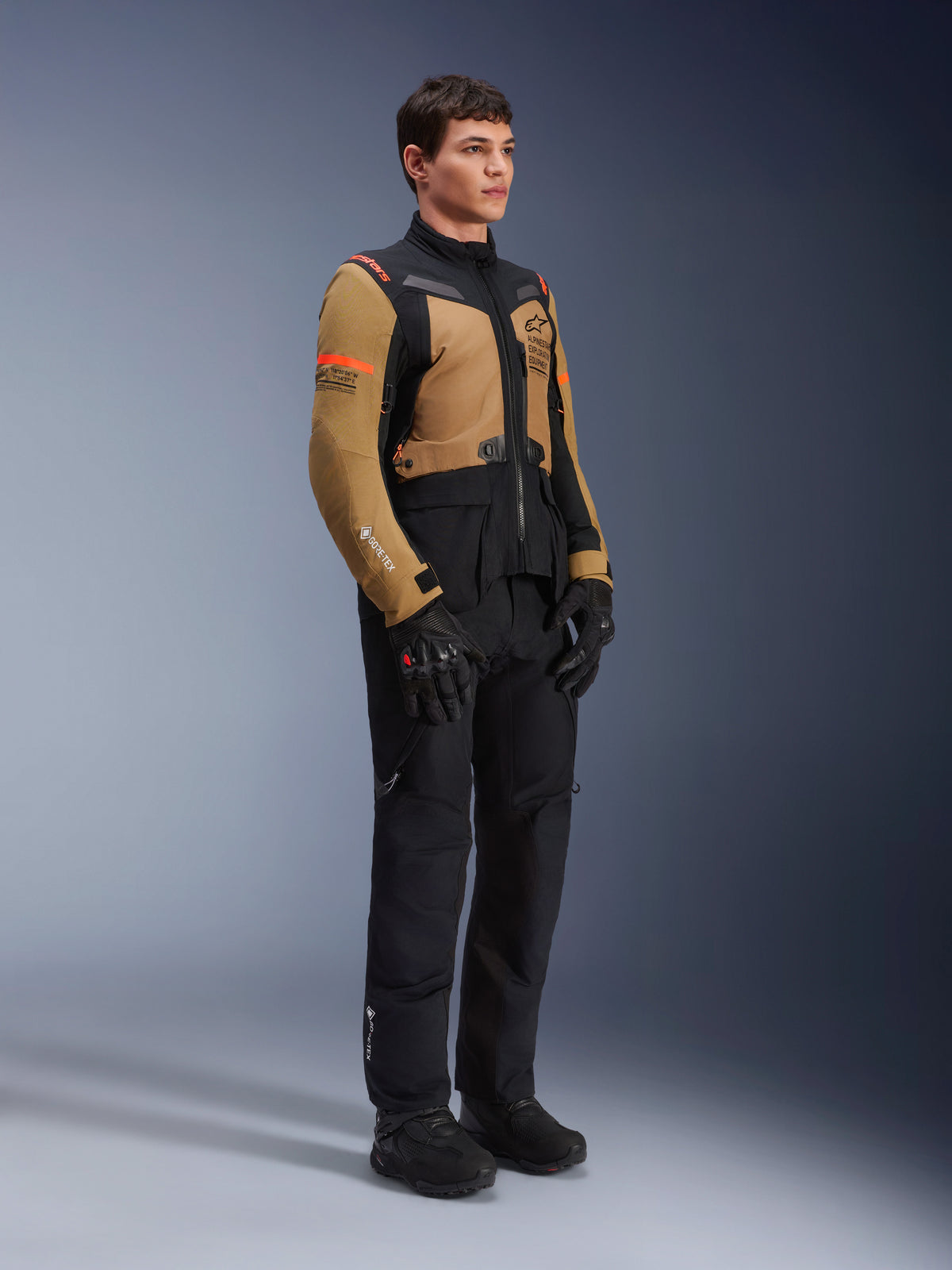 Alpinestars ST-7 2L GORE-TEX Jas, motorjas, Gobi bruin en zwart, gedragen door een model met bijpassende zwarte broek en handschoenen, voorzien van bruine panelen op de borst en mouwen, zwarte technische accenten en oranje logo's, adventure-touring motorkleding.