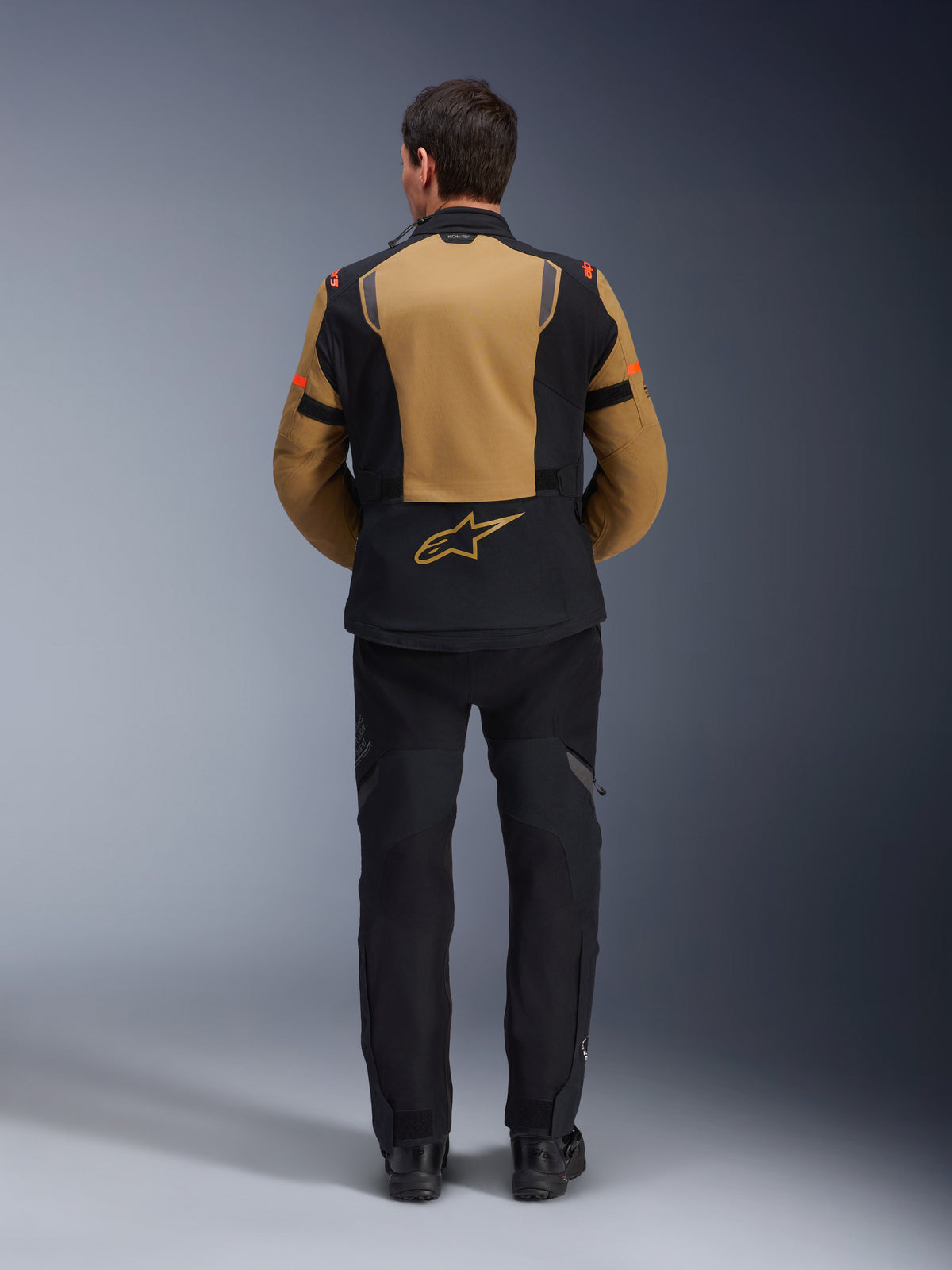 Alpinestars ST-7 2L GORE-TEX Jas, adventure motorjas, Gobi Brown en zwart, achteraanzicht met ergonomische panelen en Alpinestars-logo, gedragen met zwarte motorbroek en laarzen, waterdichte touringuitrusting
