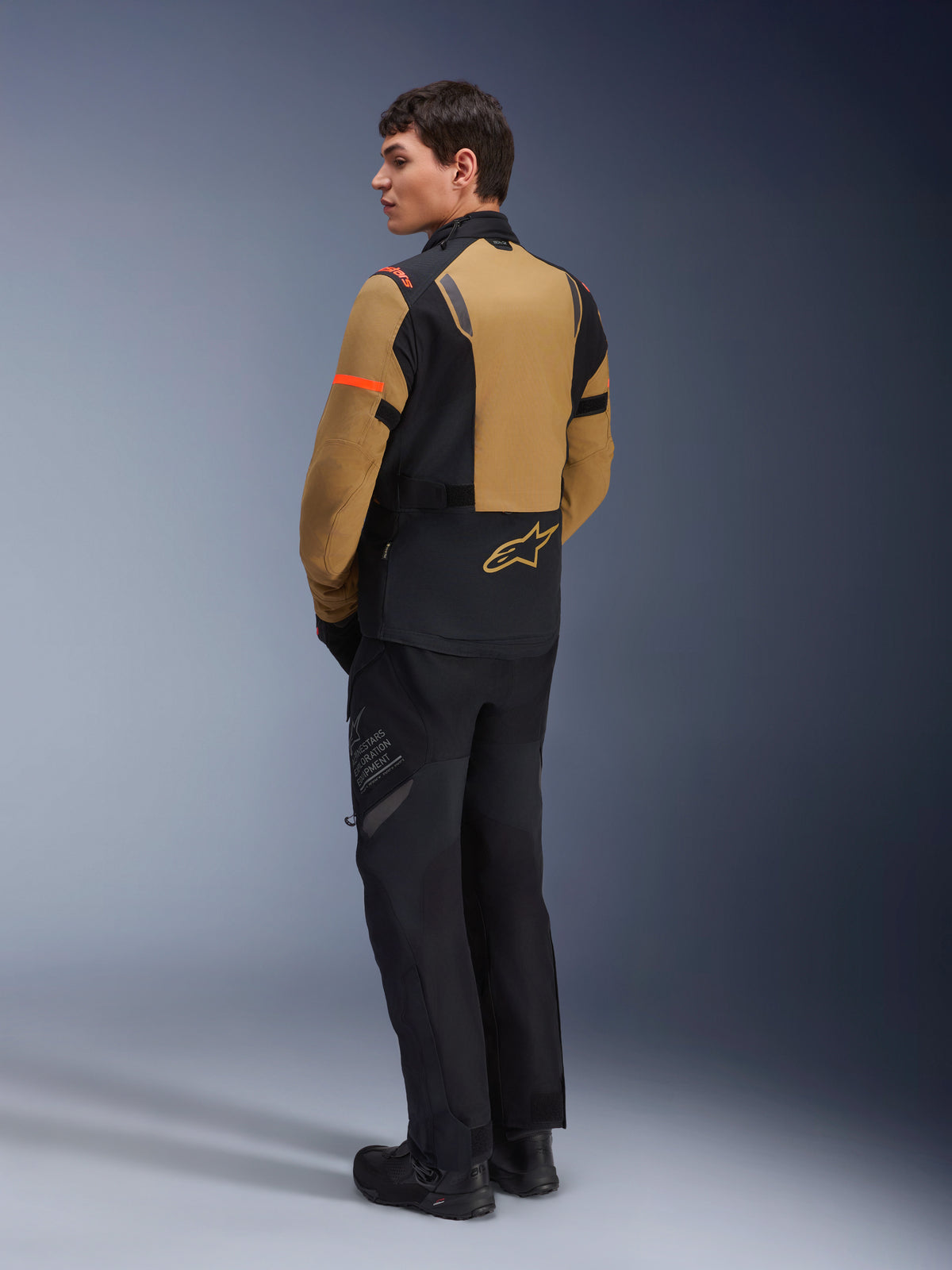 Alpinestars ST-7 2L GORE-TEX jas, adventure motorjas, Gobi bruin en zwart, achteraanzicht op een model met bijpassende motorbroek en laarzen, voorzien van oranje accenten op de mouwen, waterdichte gelamineerde schaal en Alpinestars-logo, ontworpen voor touring en rijden op de weg