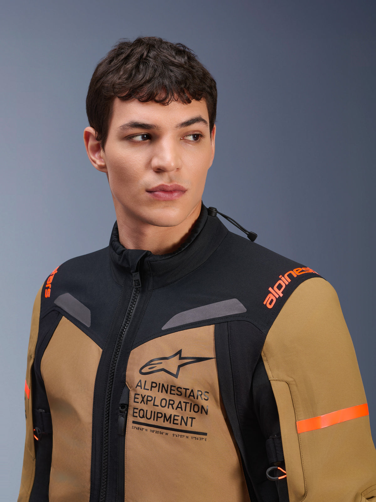 Alpinestars ST-7 2L GORE-TEX Jas, Gobi Brown en zwart, gedragen door een mannelijk model in een studio-omgeving, met prominente 'ALPINESTARS EXPLORATION EQUIPMENT' branding op de borst, oranje schouderlogo's en technische waterdichte textielconstructie voor adventure rijden