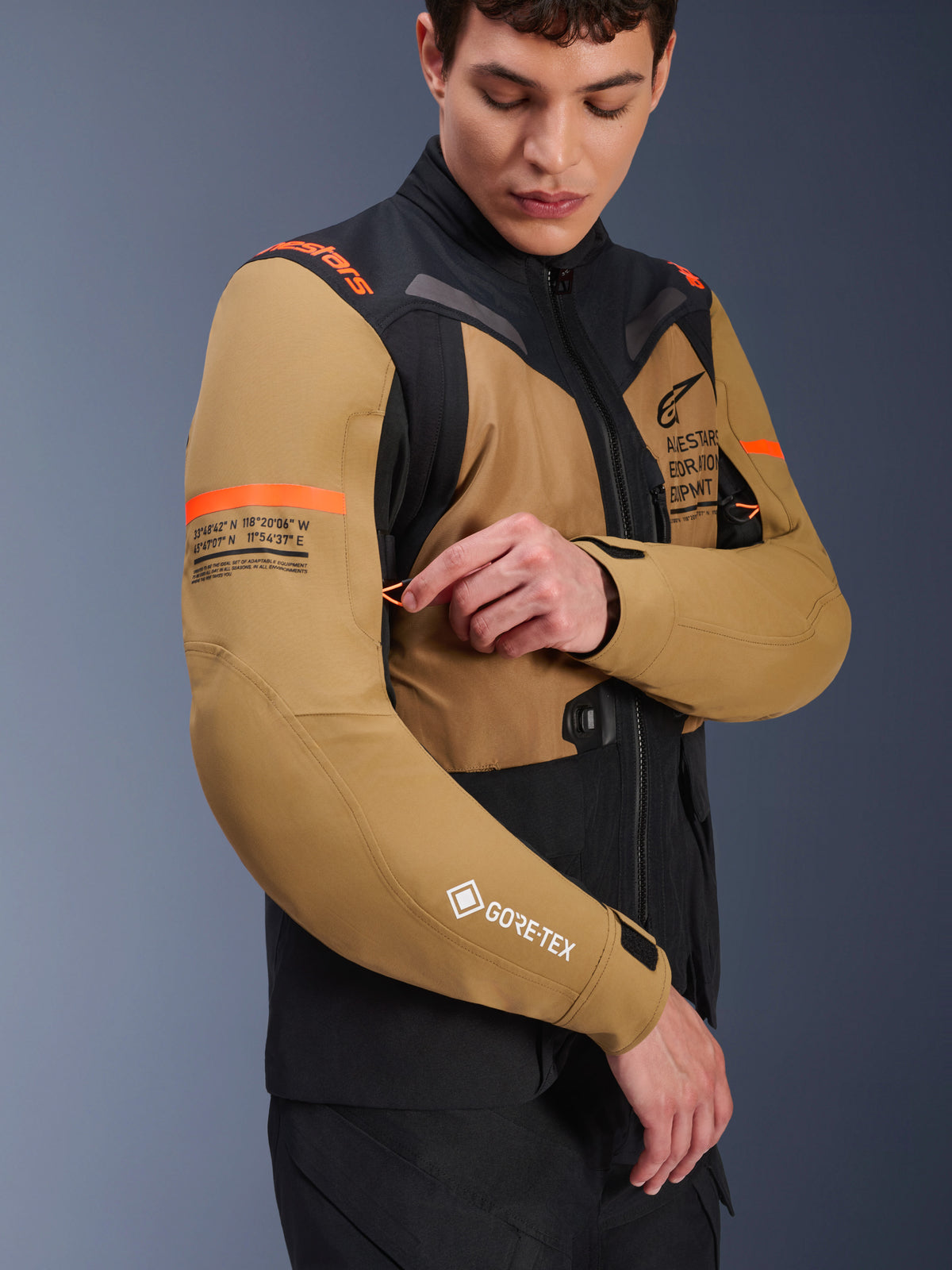 Alpinestars ST-7 2L GORE-TEX Jas in Gobi Brown en zwart, adventure-touring motorjas gedragen door een model die een ventilatieopening op de mouw aanpast, voorzien van oranje accenten, coördinatengrafieken en GORE-TEX-logo
