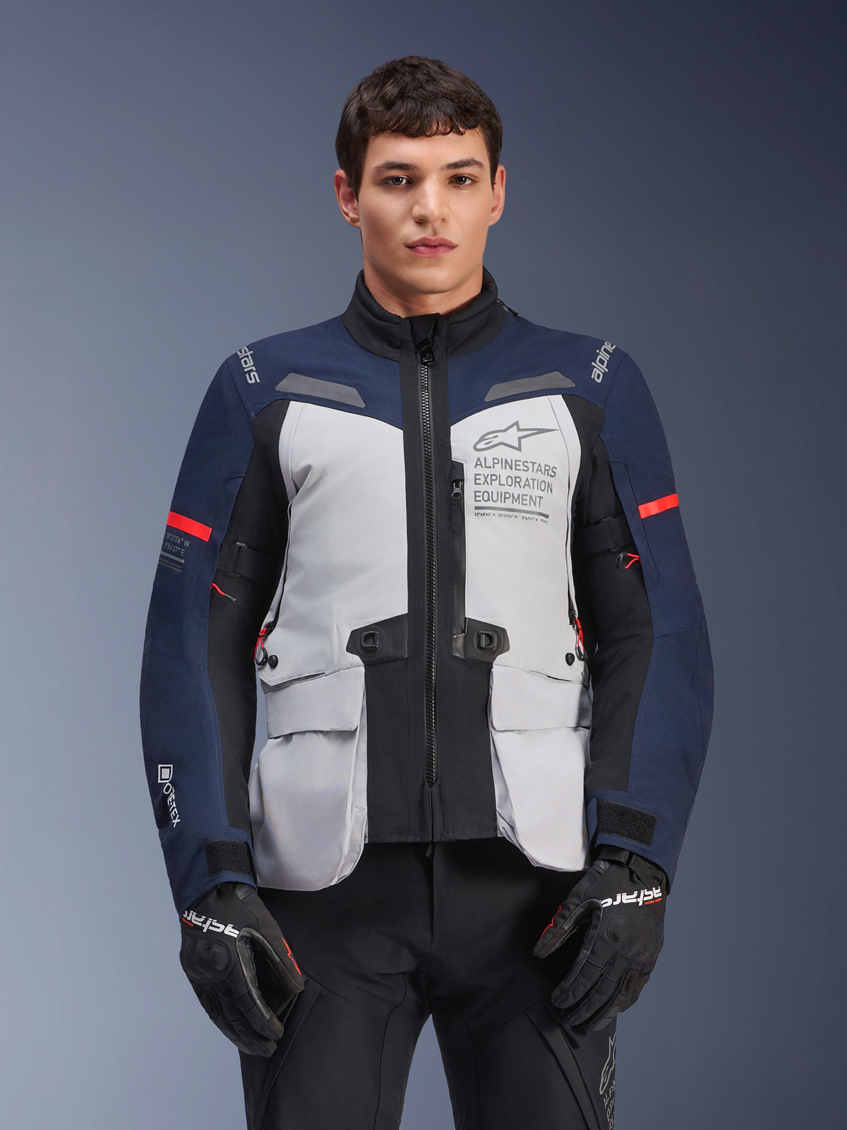 Alpinestars ST-7 2L GORE-TEX jas, motor adventure touringjas, ijsgrijs en donkerblauw met rode accenten, waterdicht en ademend, gedragen door een model met zwarte handschoenen, voorzien van Alpinestars Exploration Equipment-branding op de borst