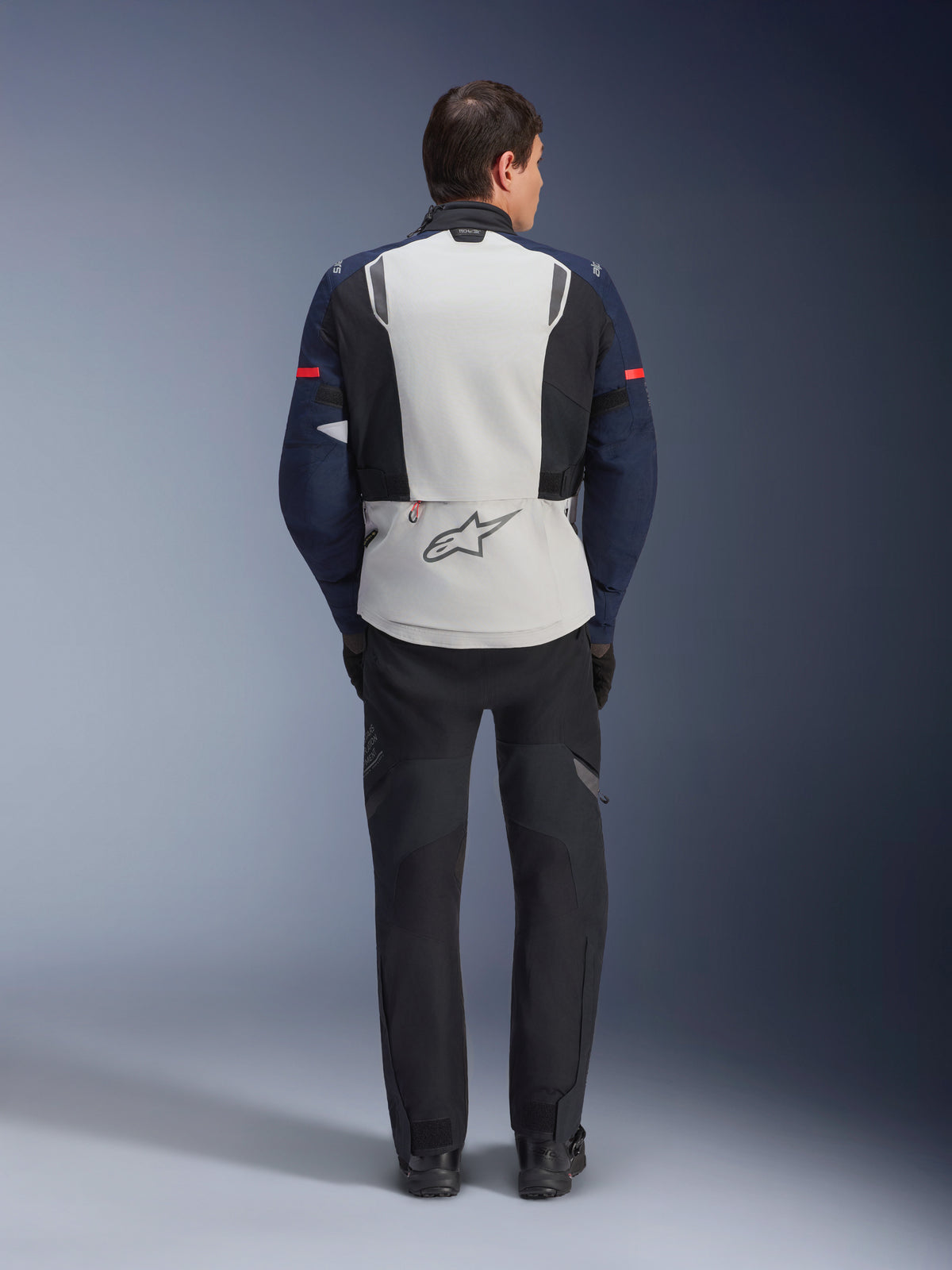 Alpinestars ST-7 2L GORE-TEX Jas, achteraanzicht van een motorrijder die een adventure touring motorjas draagt in Ice Gray, Dark Blue en zwart, voorzien van een groot rugpaneel met Alpinestars-logo en bijpassende zwarte broek, ontworpen voor alle weersomstandigheden.