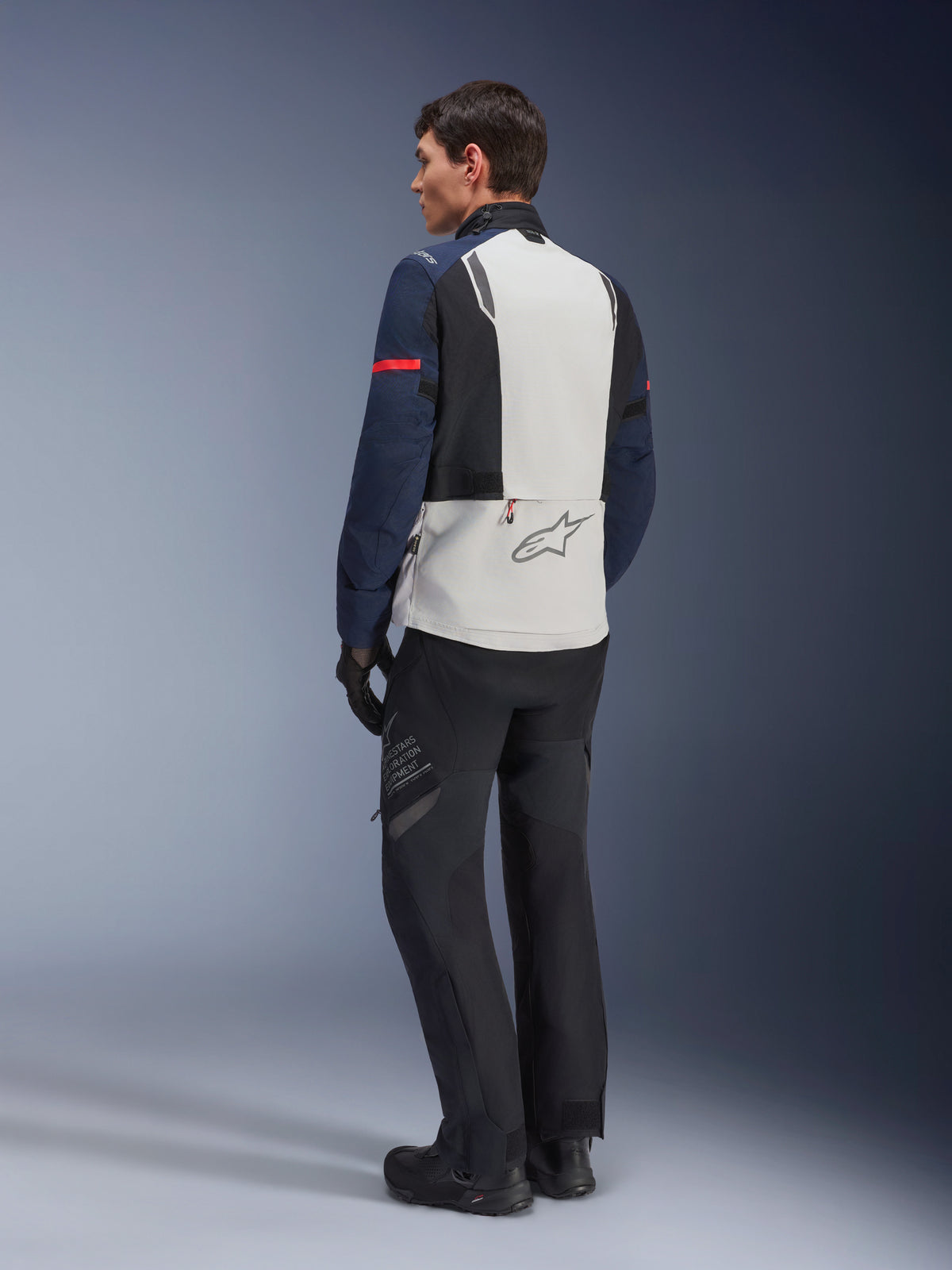 Model draagt Alpinestars ST-7 2L GORE-TEX Jas in Ice Gray, donkerblauw en zwart, achteraanzicht, lichtgrijs rugpaneel met Alpinestars-logo, donkerblauwe mouwen met rode streep, gecombineerd met zwarte adventure motorbroek en laarzen, motor touring uitrusting
