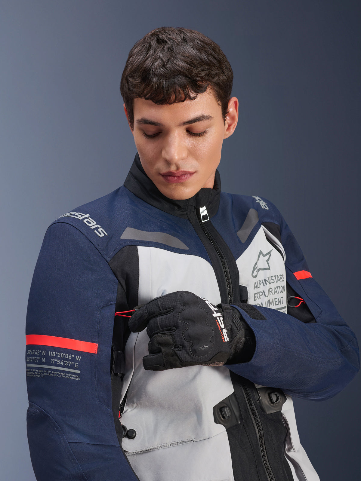 Alpinestars ST-7 2L GORE-TEX jas, Ice Gray, donkerblauw en zwart met oranje accenten, gedragen door een man die een detail op de mouw aanpast, voorzien van 'Alpinestars Exploration Equipment' branding en geografische coördinaten, professionele adventure motorrijuitrusting