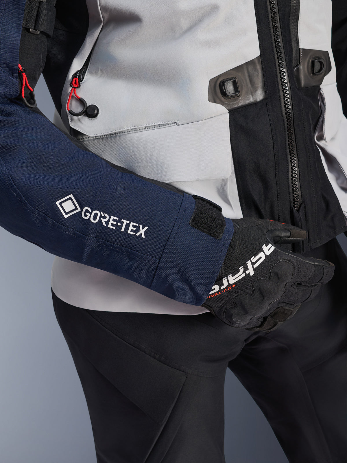 Alpinestars ST-7 2L GORE-TEX Jas, close-up van donkerblauwe mouw met wit GORE-TEX logo, ijsgrijs torso met zwarte details en rode koordaccenten, gedragen met zwarte Alpinestars handschoenen en broek, adventure touring motorkleding