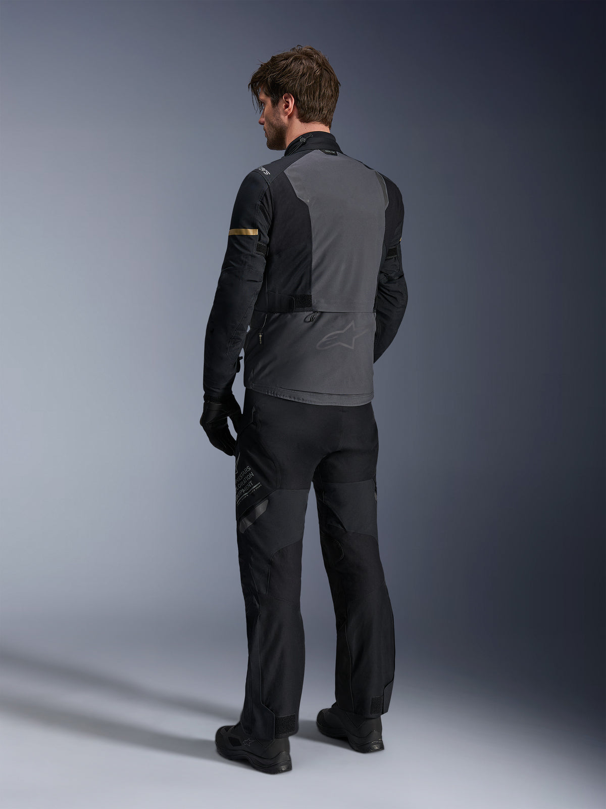 Alpinestars ST-7 2L GORE-TEX Jacket, Deep Gray and Black with Dark Khaki accents, rear view of a rider wearing the jacket and matching motorcycle pants, adventure touring gear with waterproof laminated construction