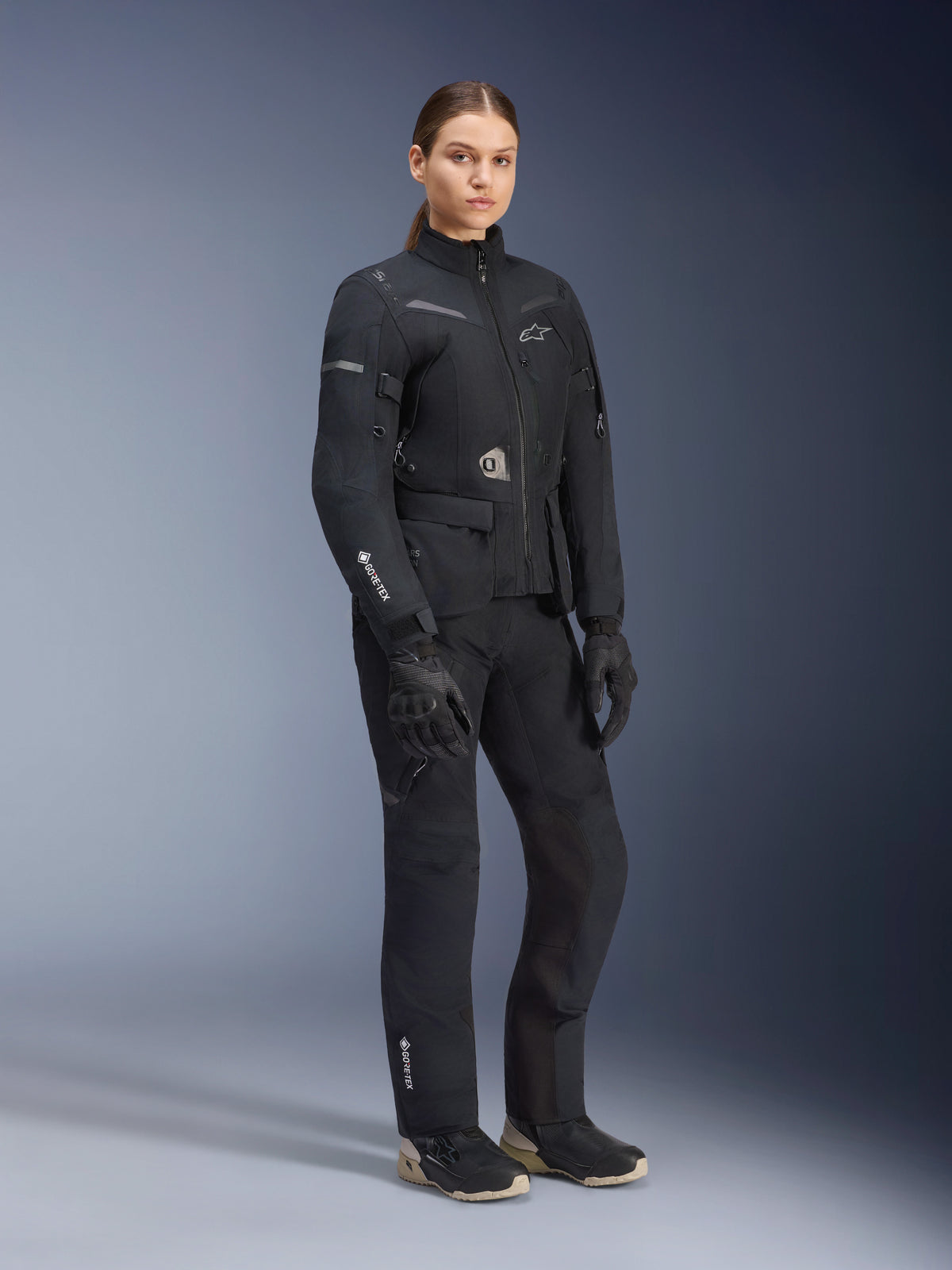 Mannequin femme portant une tenue d'aventure touring Alpinestars Stella ST-7 2L GORE-TEX, veste et pantalon noir et gris foncé, arborant le logo GORE-TEX sur la manche et la jambe, associée à des gants noirs et des bottes de moto, équipement de moto professionnel conçu pour le touring toutes saisons.