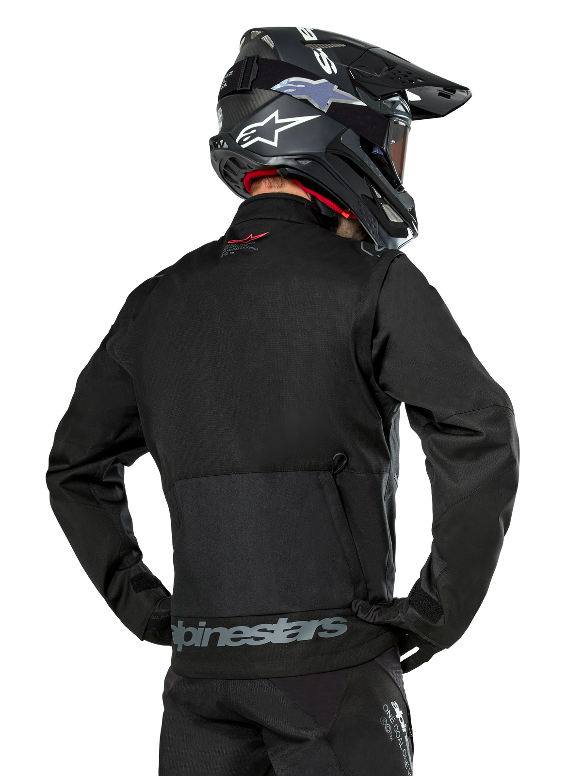 Vue arrière d'un pilote portant la veste Alpinestars Pro-Dura en noir, veste d'enduro durable en textile ripstop, dotée d'une grande poche cargo arrière avec marquage Alpinestars gris sur le bas du dos, associée à un casque de motocross noir.