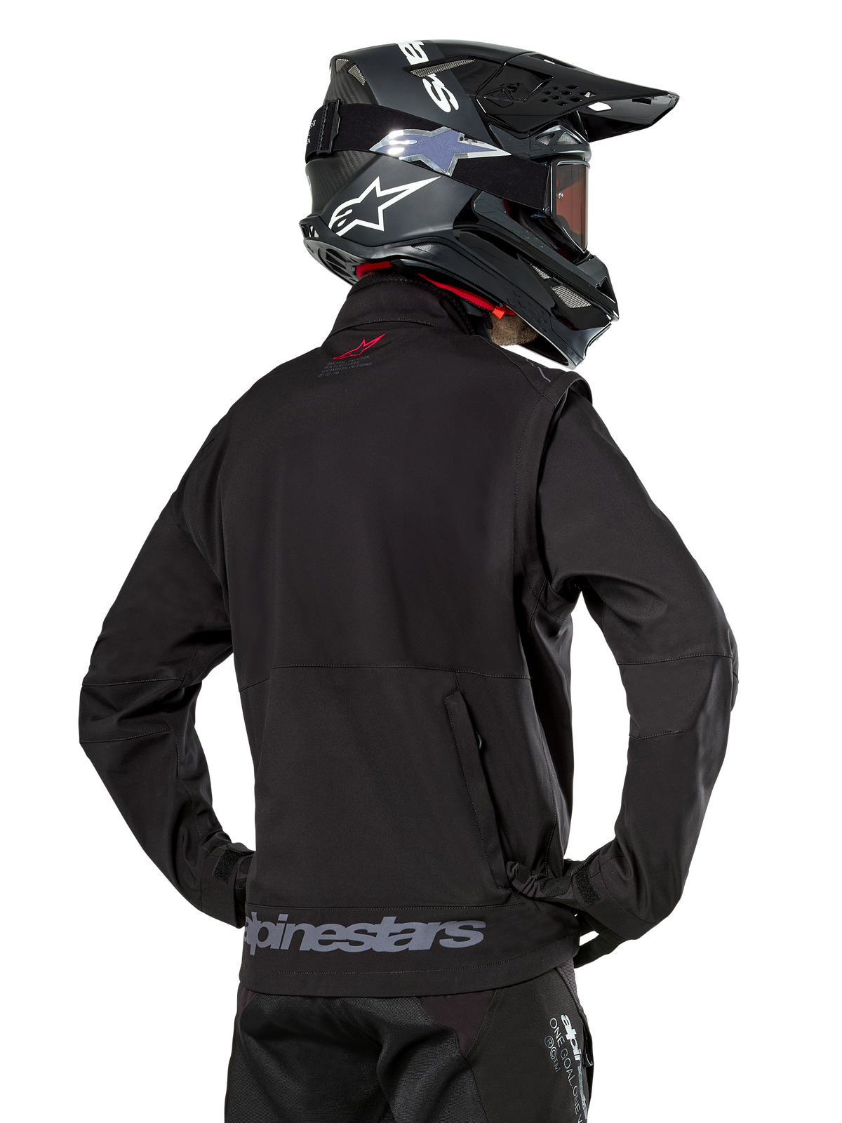 Alpinestars Lite-Dura Softshell-Jacke, Schwarz, Rückansicht getragen von einem Fahrer, mit grauem Alpinestars-Branding am hinteren Saum und einem kleinen roten Logo am Kragen, kombiniert mit einem schwarzen Motocross-Helm, entwickelt für Enduro- und Offroad-Fahrten.