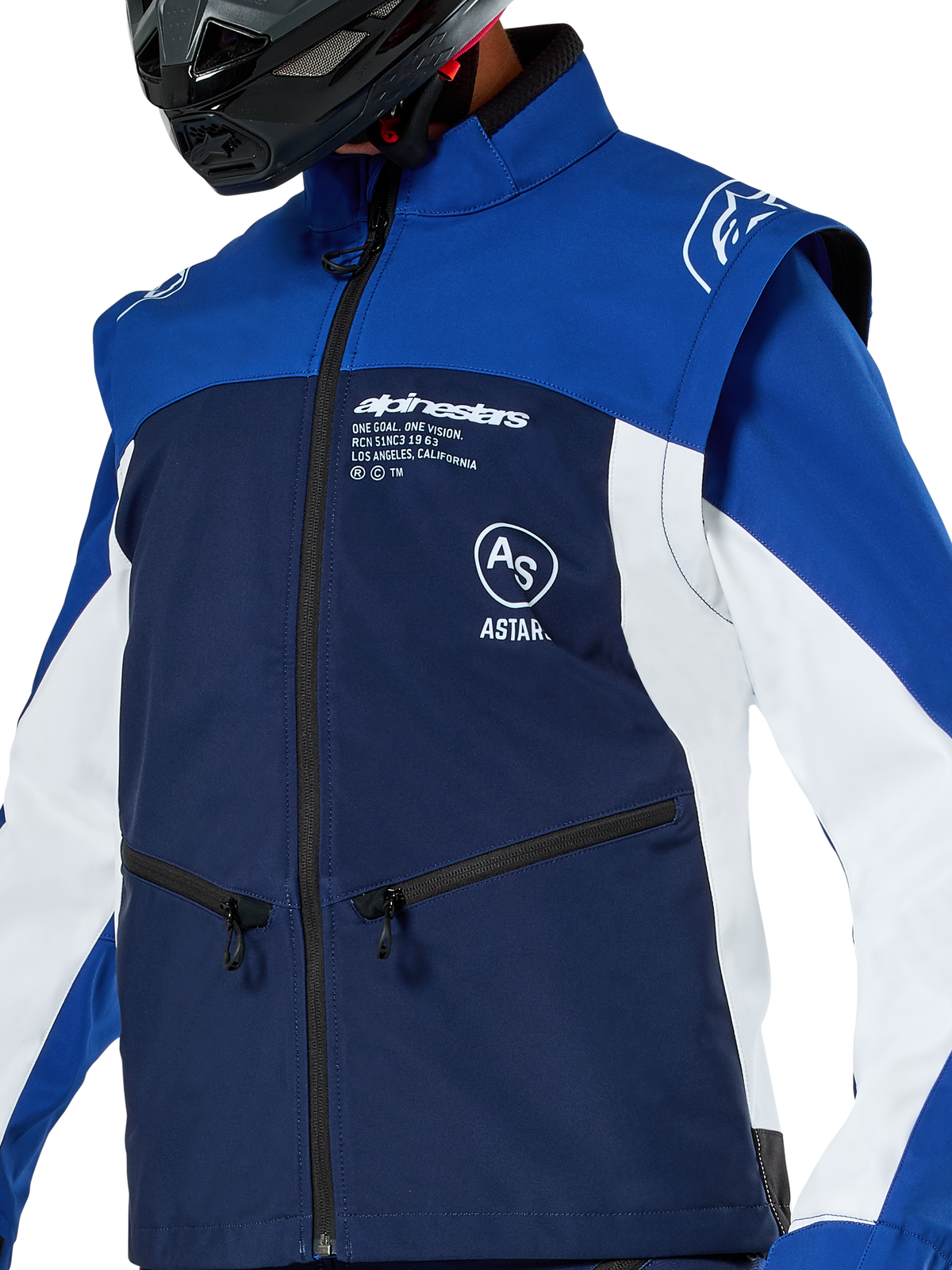 Alpinestars Lite-Dura Softshell-Jacke, Motocross- und Enduro-Jacke, Dunkelblau, Hellblau und Weiß, mit weißem Alpinestars-Branding auf der Brust und Reißverschlusstaschen, getragen mit einem schwarzen Helm, strapazierfähige Softshell-Konstruktion mit abnehmbaren Ärmeln für Offroad-Fahrten.