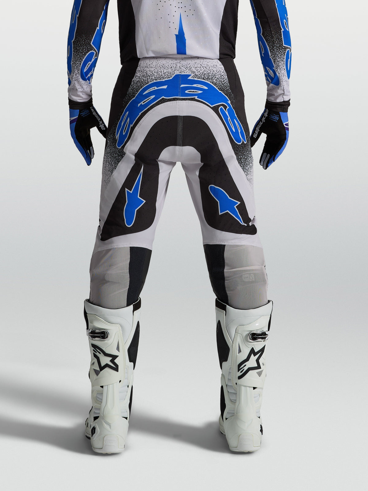 Alpinestars Supertech Hiten AFD broek, herenmotorcrossbroek voor off-road racing, blauw, zwart en wit, atletische racepasvorm, van achteren gezien op een model met witte motorlaarzen.