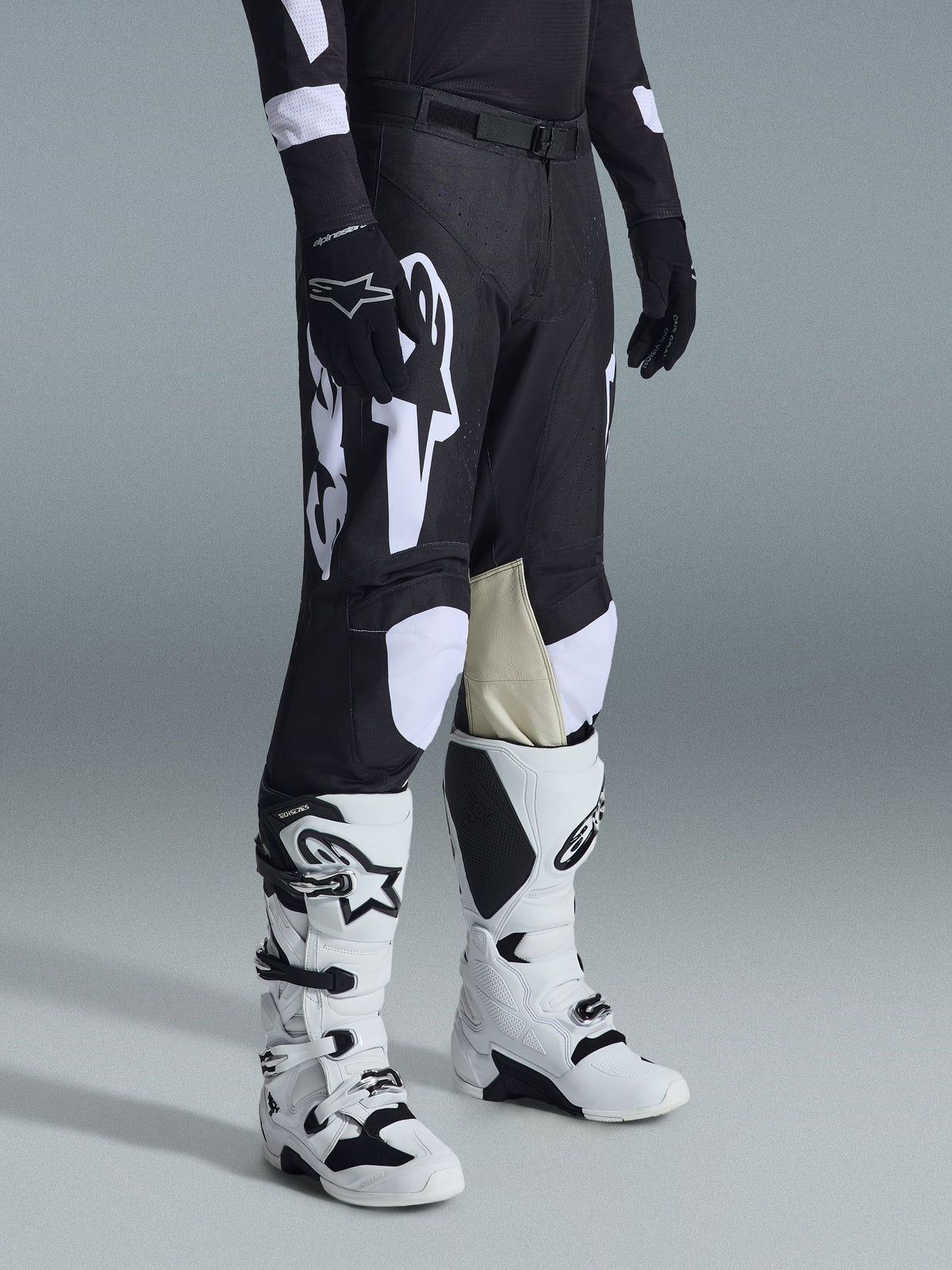 Alpinestars 2026 Racer Air Riway Hose, Schwarz-Weiße Motocross-Hose mit großen weißen Logo-Grafiken, beigen Lederbahnen an den Knieinnenseiten, getragen mit weiß-schwarzen Offroad-Stiefeln und schwarzen Handschuhen in einer Studio-Umgebung.