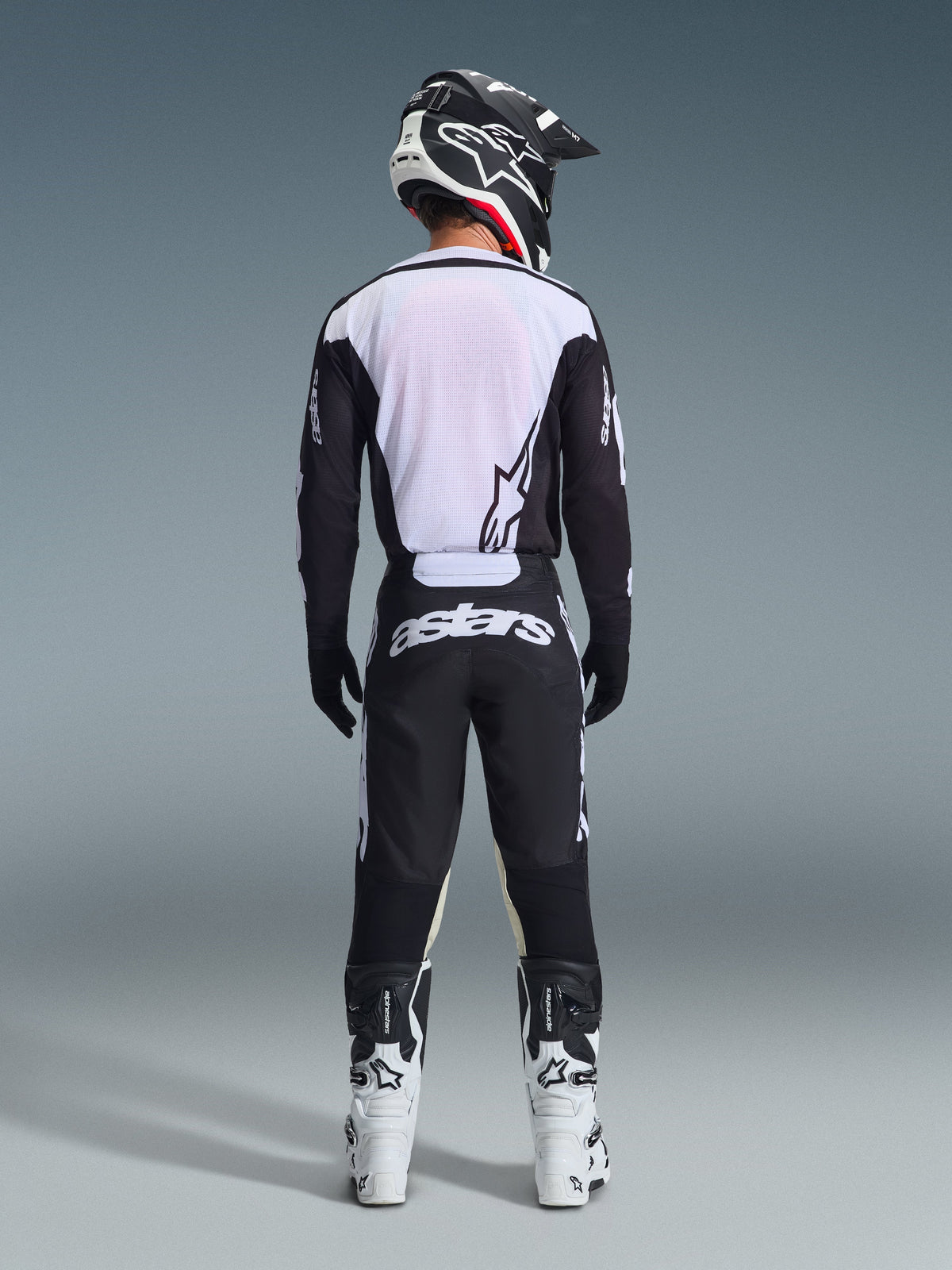Pilote de motocross vu de dos portant le pantalon Alpinestars 2026 Racer Air Riway noir et blanc, maillot à manches longues assorti, bottes de course blanches et noires et casque noir avec logo Alpinestars