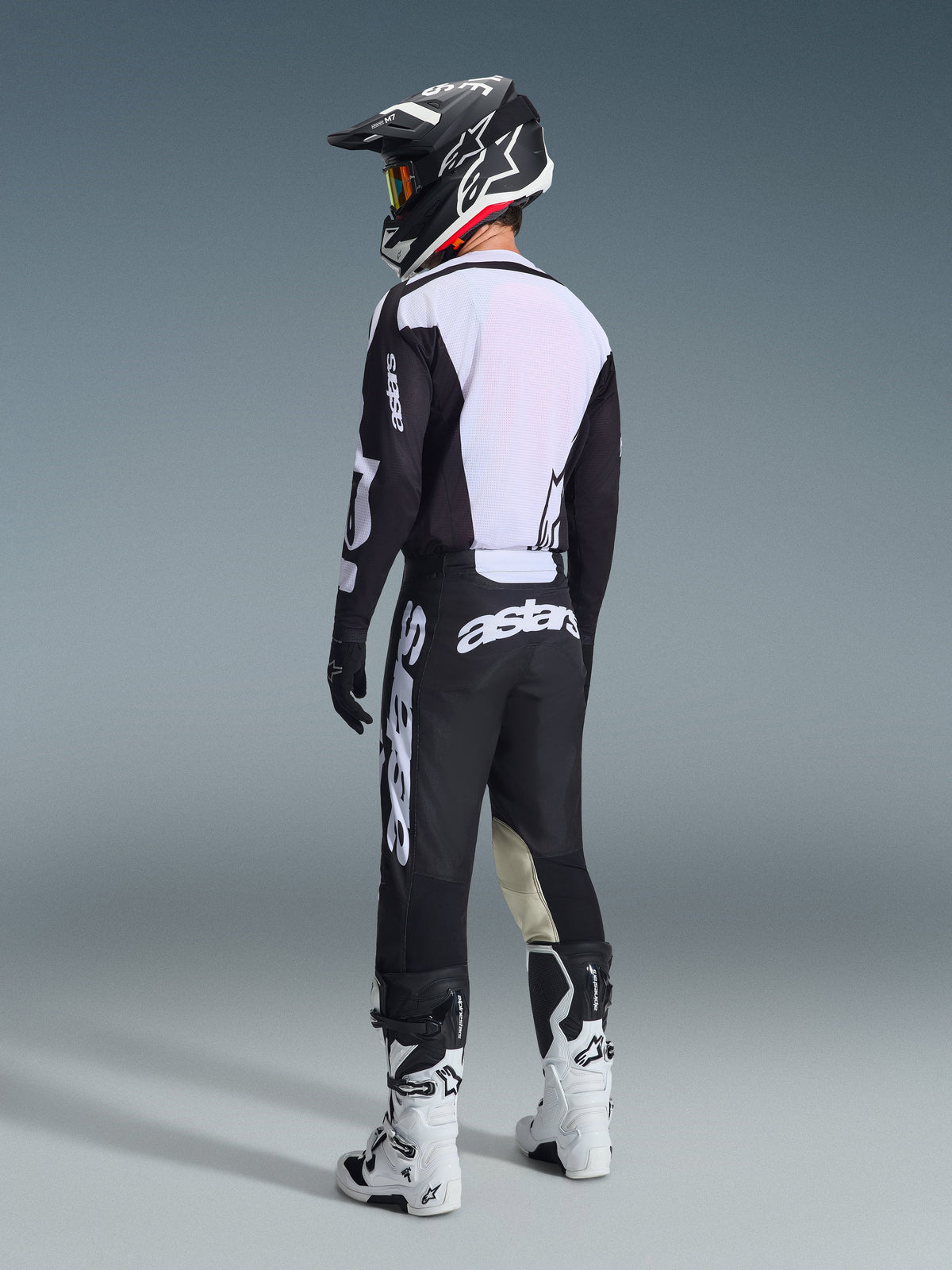 Pilote de motocross vu de dos portant le pantalon Alpinestars 2026 Racer Air Riway en noir et blanc, associé à un maillot ventilé assorti, un casque noir et blanc et des bottes de motocross blanches, avec un marquage blanc 'astars' distinctif sur l'assise.