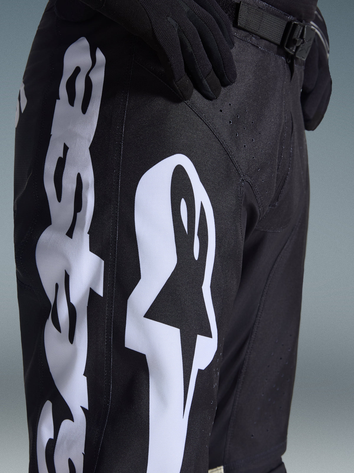 Alpinestars 2026 Racer Air Riway Hose, schwarze Motocross-Hose mit markantem weißem Alpinestars-Logo auf dem Oberschenkel, Nahaufnahme von technischem Gewebe und verstellbarem Taillenriemen, getragen mit schwarzen Handschuhen für den Offroad-Sport