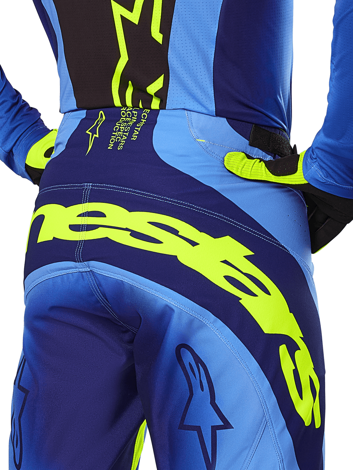 Alpinestars 2025 Techstar Melt crossbroek, motorcrossbroek, geel fluo en blauw, close-up van het achteraanzicht met opvallende gele Alpinestars-branding op het zitvlak, gedragen met bijpassende blauwe jersey en geel-fluorescerende handschoenen, ontworpen voor off-road racing.
