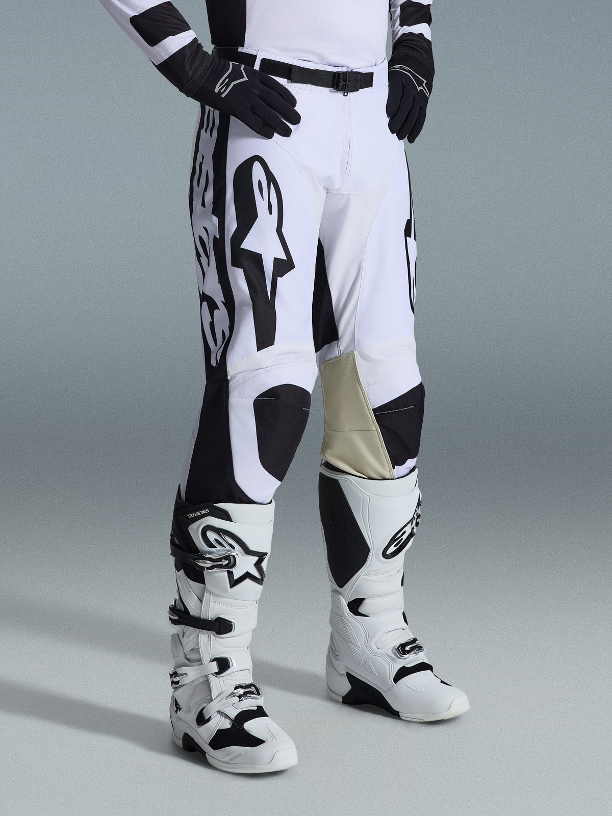 Alpinestars 2026 Racer Riway Pants, motorcrossbroek, wit en zwart, voorzien van opvallende zwarte Alpinestars-graphics en logo's, zandkleurige lederen knieverstevigingen aan de binnenkant, gedragen met wit-zwarte motorcrosslaarzen en zwarte handschoenen, professionele race-uitrusting voor off-road rijden
