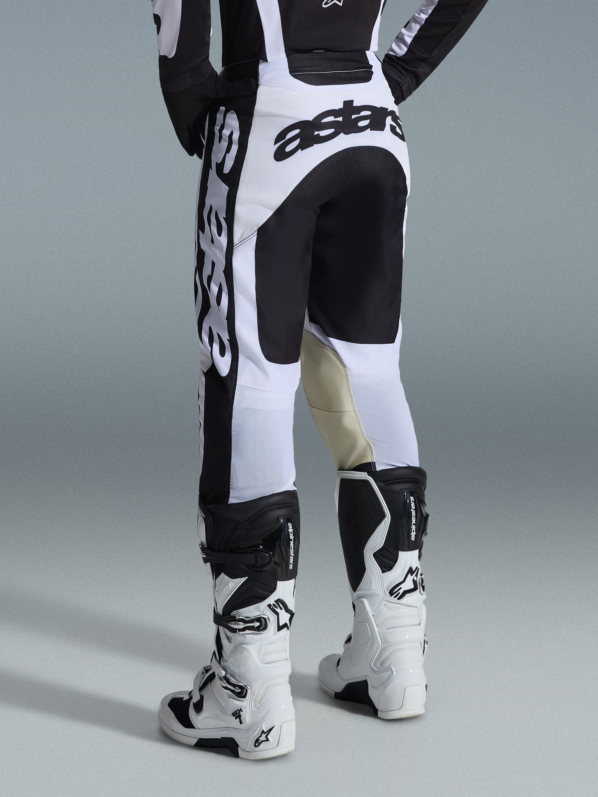 Alpinestars 2026 Racer Riway Pants, motorcrossbroek, wit en zwart, achteraanzicht met opvallende zwarte branding op het zitvlak en de zijkanten, beige geitenleren panelen aan de binnenkant van de knieën, gedragen met bijpassende wit-zwarte motorcrosslaarzen