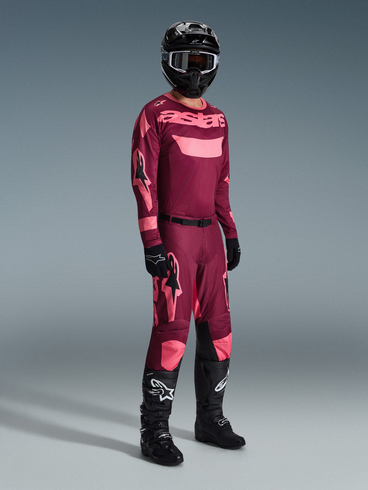 Motocross-Fahrer in kompletter Rennausrüstung, Alpinestars 2026 Racer Riway Trikot in Dark Red Coral mit pinkfarbenen Akzenten, passende dunkelrote Hose mit pinkfarbenen Logos, schwarzer Helm mit weiß umrandeter Brille, schwarze Handschuhe und schwarze Stiefel mit weißen Logos, koordiniertes Outfit für Offroad-Rennen
